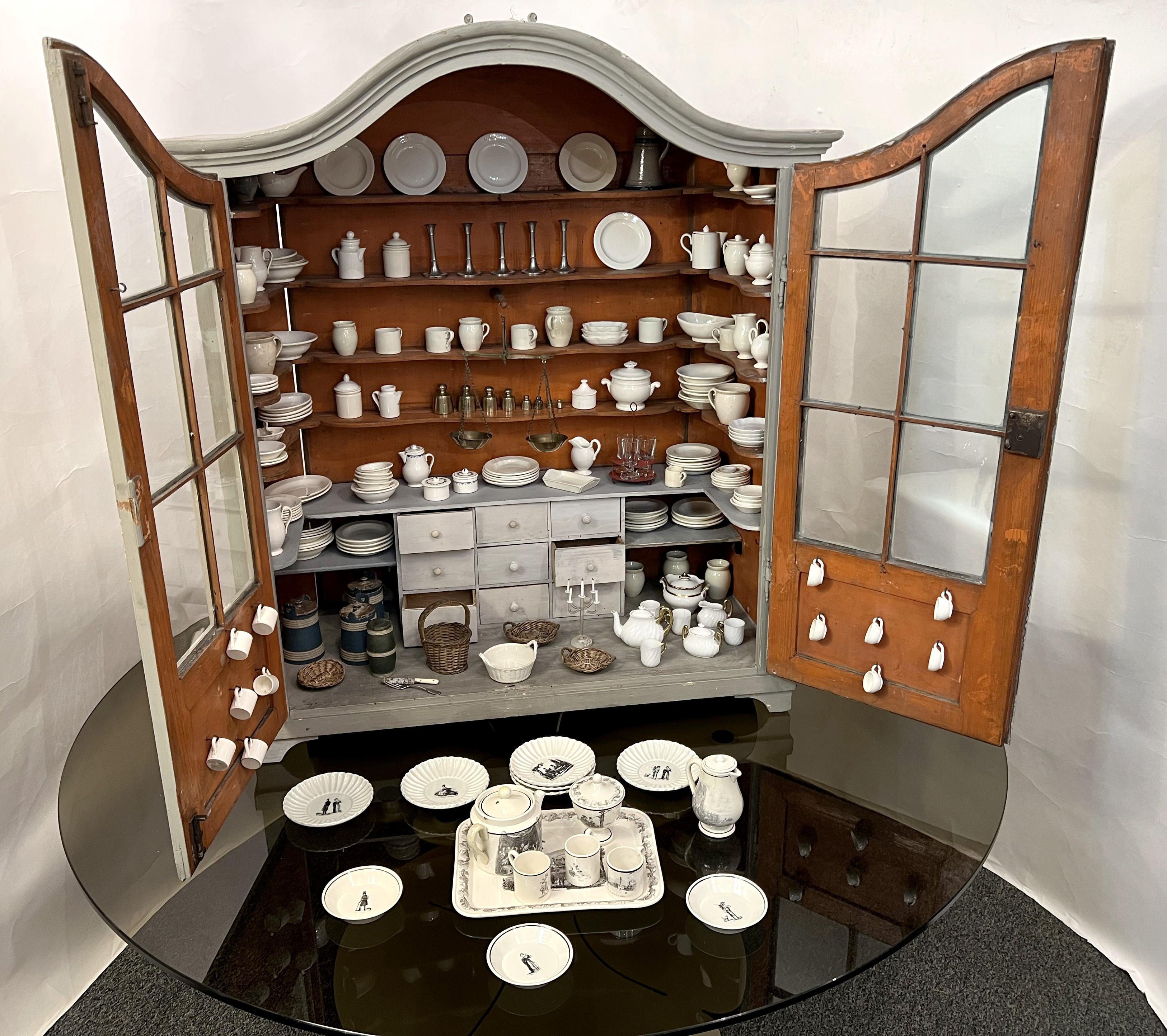 Doll's vaisselier in grey lacquered wood opening by two partially glazed doors. Entirely furnished with porcelain and fine earthenware dishes, including a large number of plates, covered pots, pitchers, tea service, ... glassware, a scale and copper
