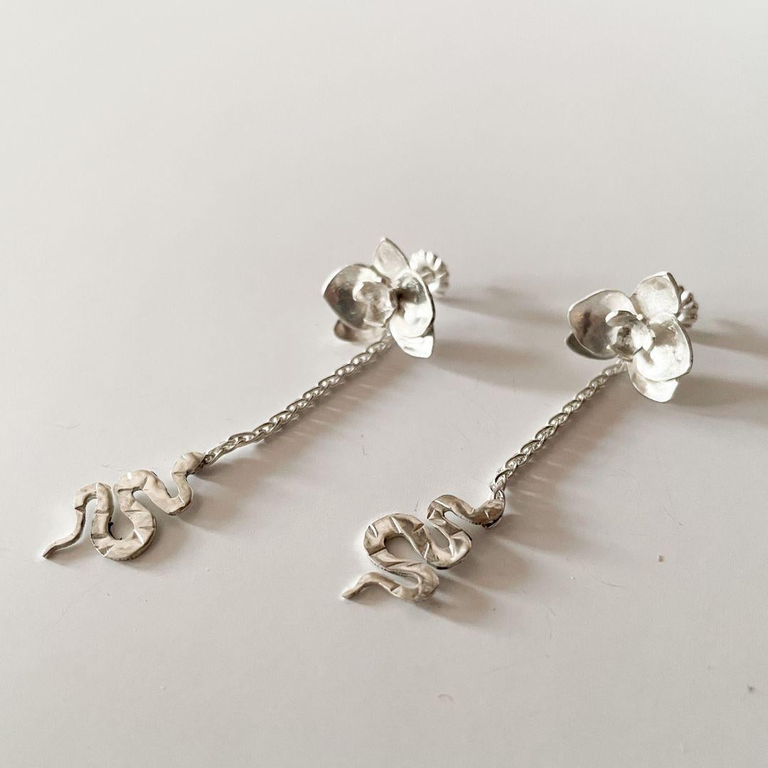 Diese handgefertigten Ohrringe aus 925er Sterlingsilber feiern die zarte Kraft der Verwandlung. An der Lasche ruht eine fein modellierte Orchidee, von der eine zierliche, kleine Schlange an einer dünnen Kette herabhängt. Die Kombination aus Blume