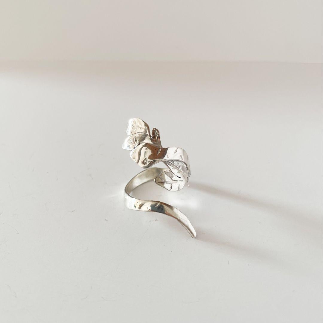 Dieser handgefertigte Ring aus 925er Sterlingsilber zeigt eine gewundene Schlange, die sich zart zwischen den skulptierten Blättern einrichtet und ein kleines Universum aus Bewegung und Symbolik an Ihrer Hand schafft. Die Schlange, uraltes Symbol