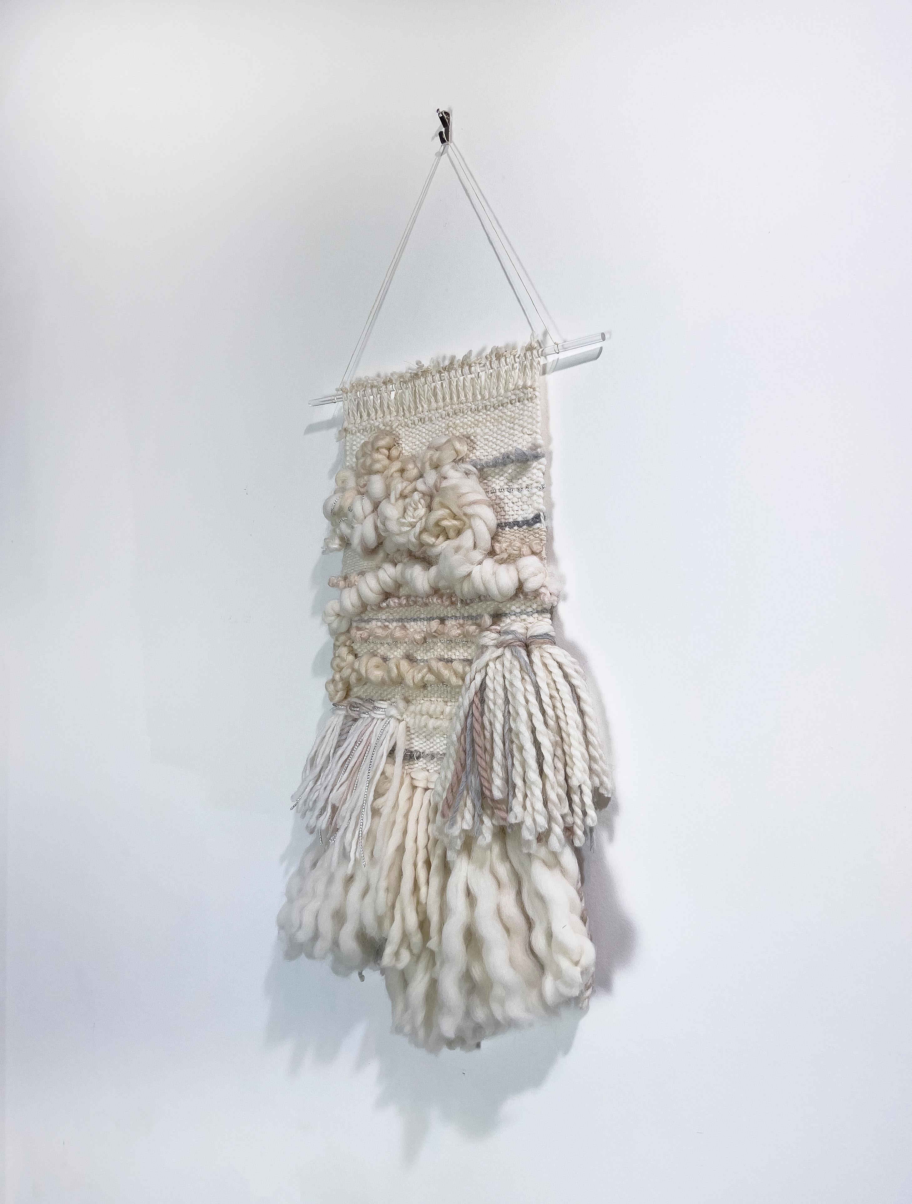 This hand-woven fiber art wall hanging by Dolores Tema is made with light white and cream-toned cotton and Alpaca and Merino wool. The weaving hangs from a clear acrylic rod, suspended by hemp twine. It is ready to hang. 

Dolores Tema studied