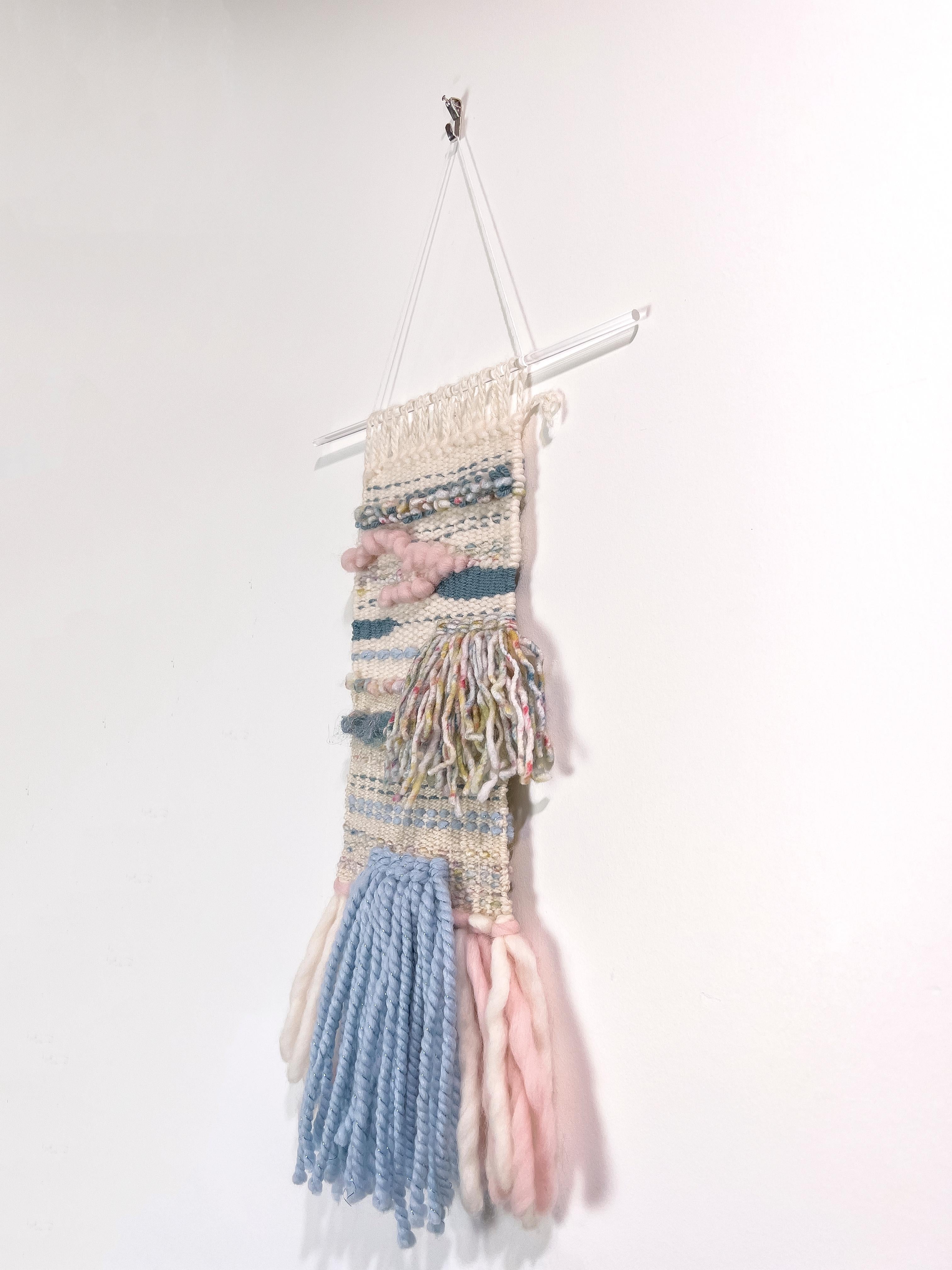 This hand-woven fiber art wall hanging by Dolores Tema is made with cotton and Alpaca and Merino wool. It features layers of woven yarn in varying colors including cream, muted pink, and muted blue and teal. The weaving hangs from a clear acrylic