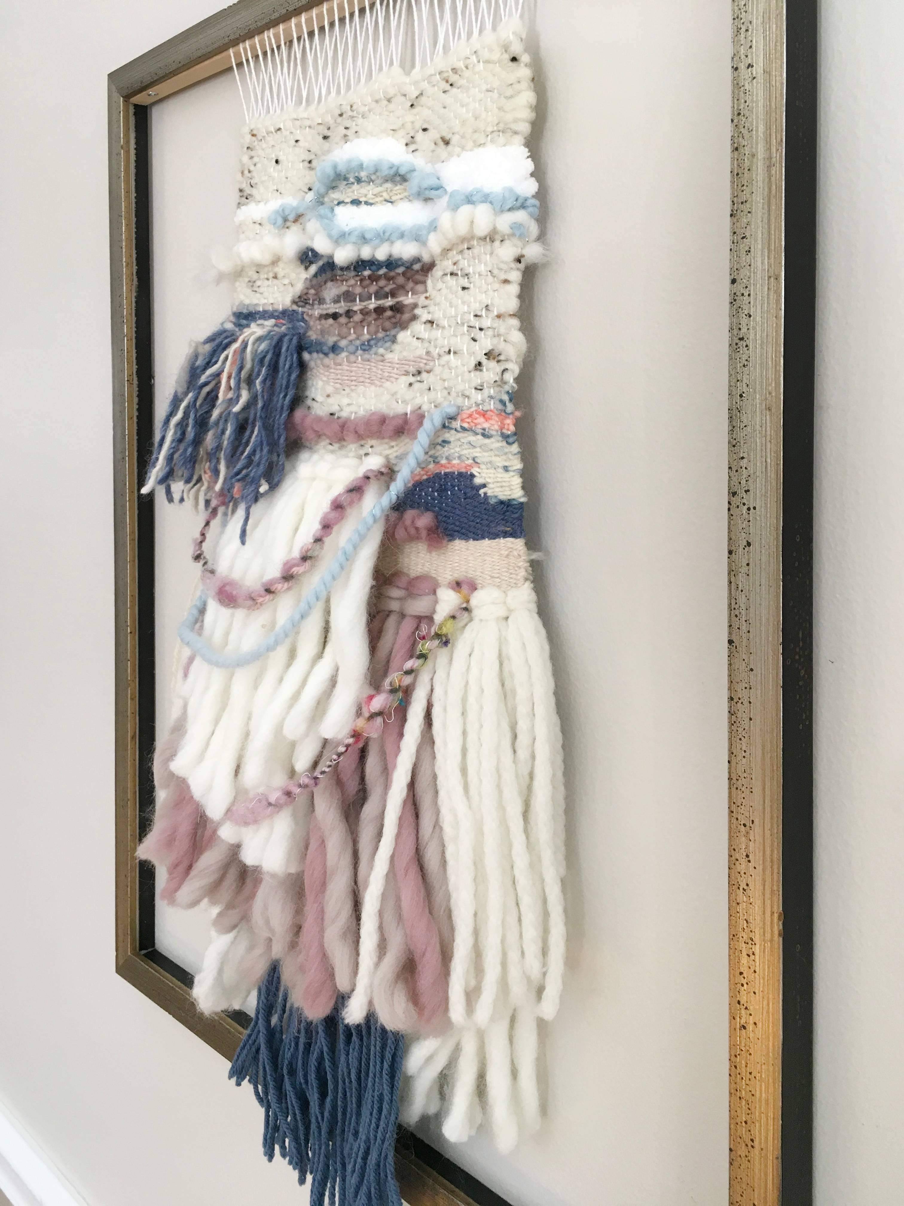 This fiber weaving by Dolores Tema is made with cotton and Merino and Alpaca wool. The weaving is framed in a distressed warm champagne frame, and is woven from the top downward, with varying colors of muted pink, beige, creme, and pink yawns woven
