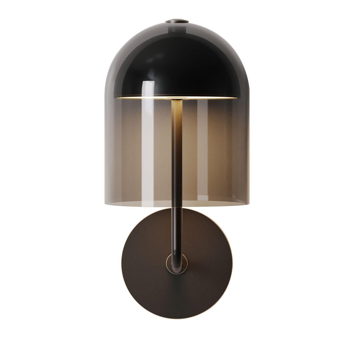 Una lampada da parete contemporanea caratterizzata da una struttura in ottone brunito scuro abbinata a un paralume a cupola in vetro grigio fumo che fornisce una luce soffusa e ambientale. Questo apparecchio di illuminazione per interni offre un