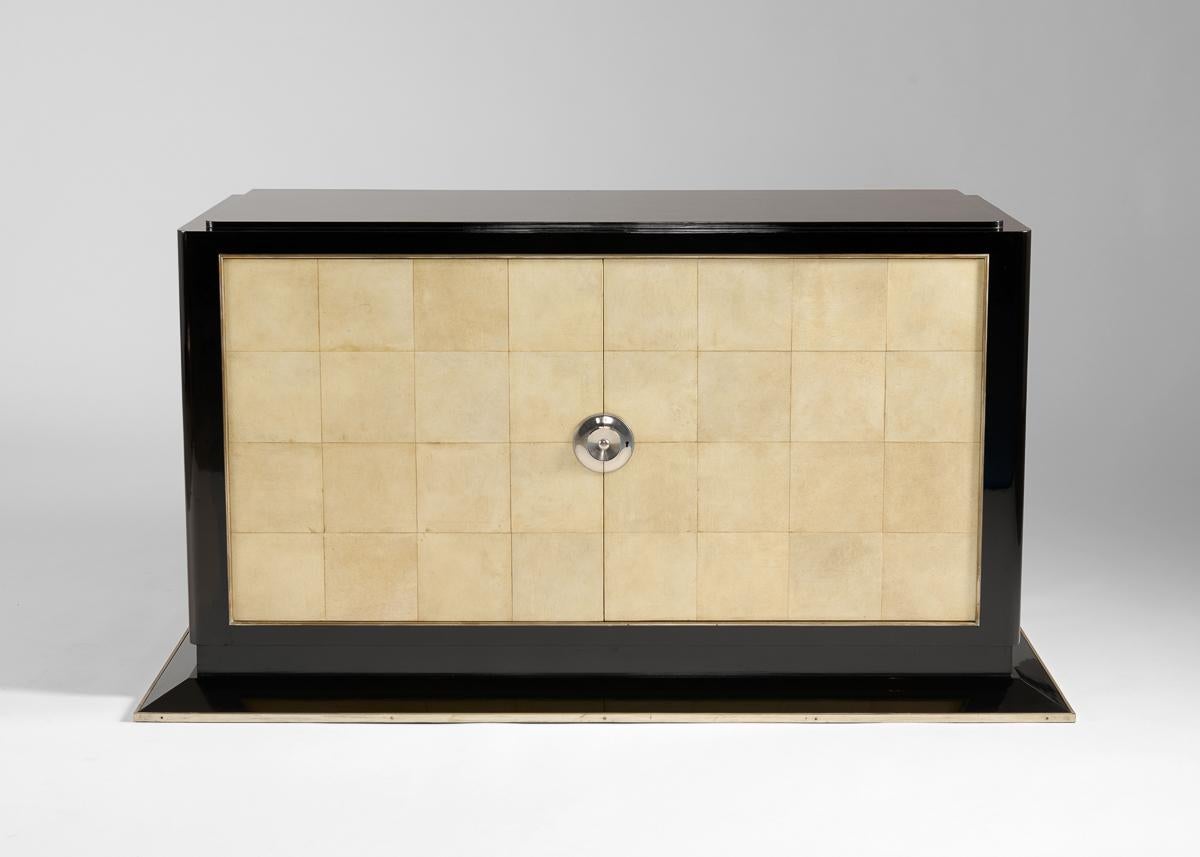 This parchment clad, black lacquered sideboard stands atop a broad base lined with nickel bronze, and possesses two doors operated by a central handle. It is representative of the duo's best, whose body of enduring designs are characterized by
