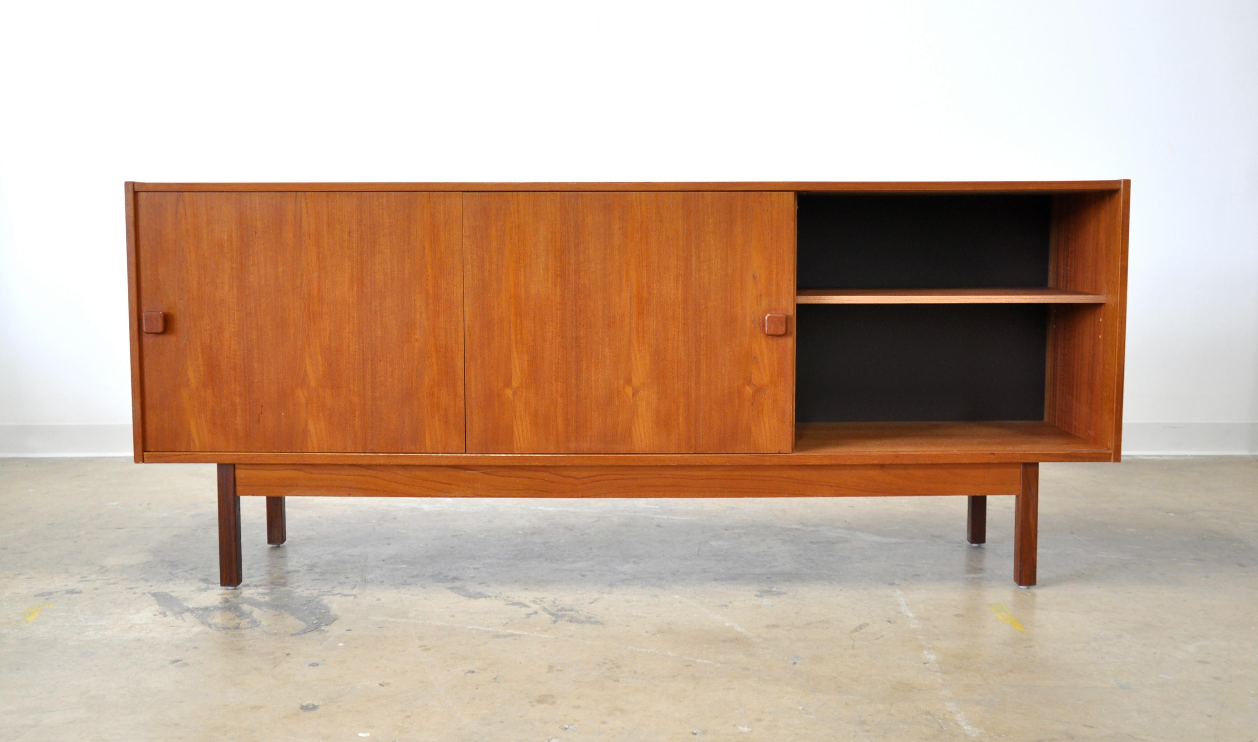 A vintage Danish Mid-Century Modern teak sideboard, made in Denmark by Domino Møbler, dating from the 1970s. The midcentury buffet features a pair of sliding cabinet doors with square sculpted handles, opening to an interior fitted with adjustable