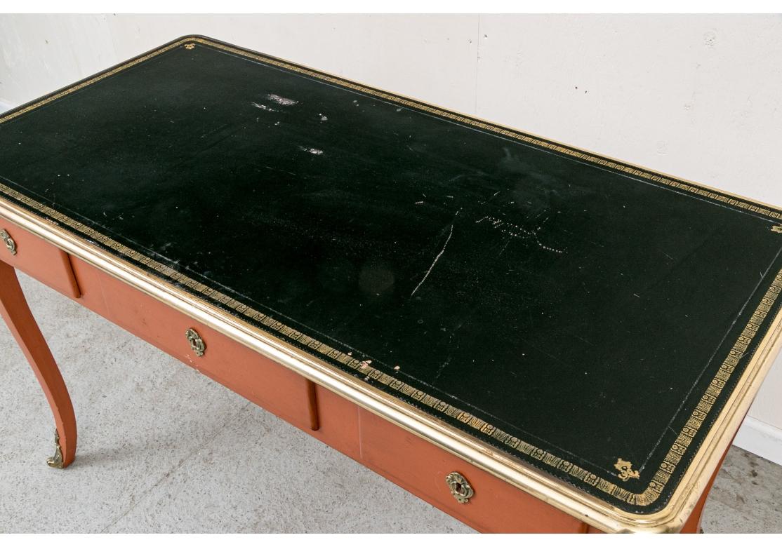 A  paint decorated writing desk with tooled black leather top, brass trim around the perimeter and brass mounts to the four legs. The desk with a larger center drawer flanked by two smaller drawers all with shield form brass escutcheons. The drawers