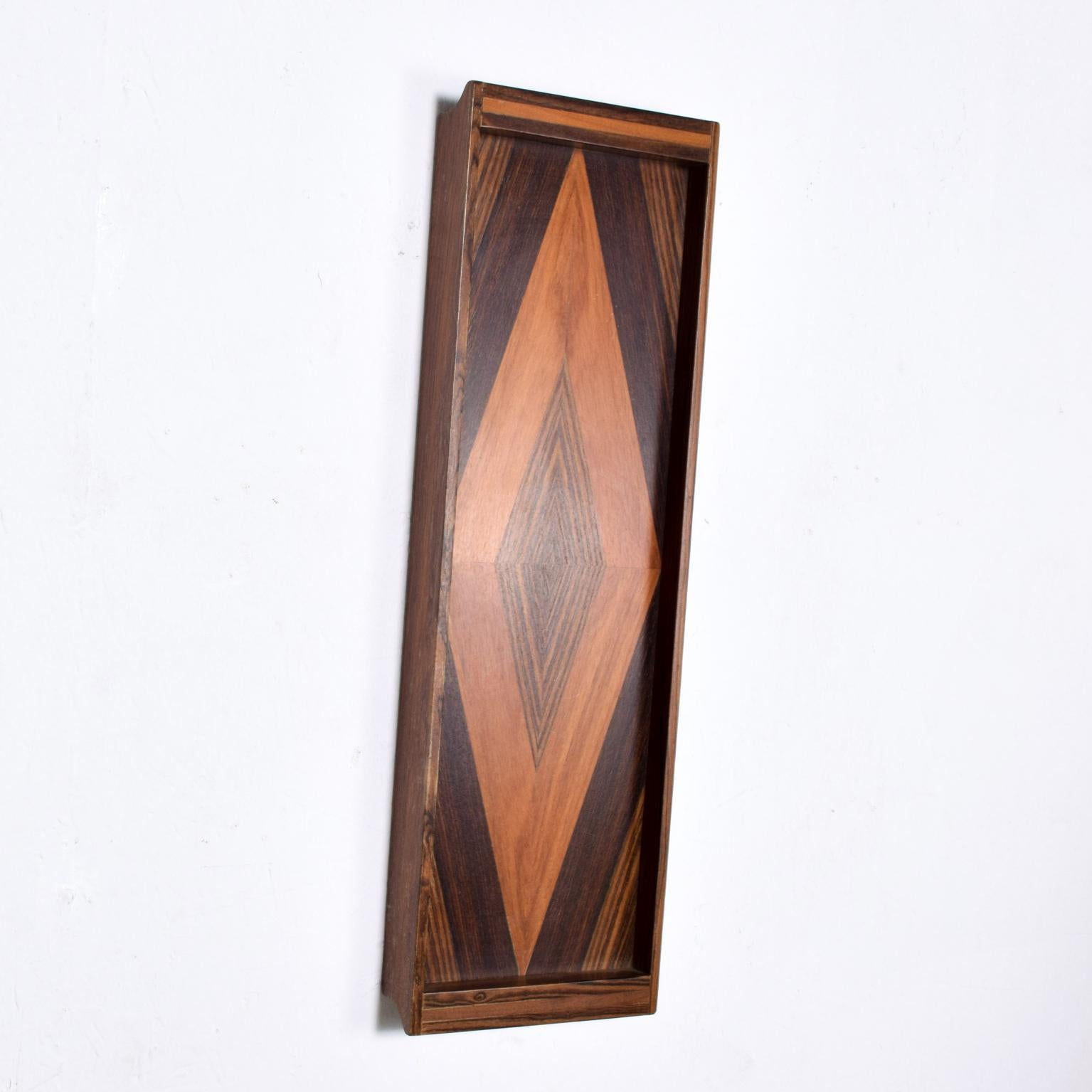 We are pleased to offer for your consideration a long and narrow service tray by Don S Shoemaker. Beautiful geometric woodwork with exotic woods. A small hole on the back of the tray allows easy hanging onto the wall. Retains label from the maker on