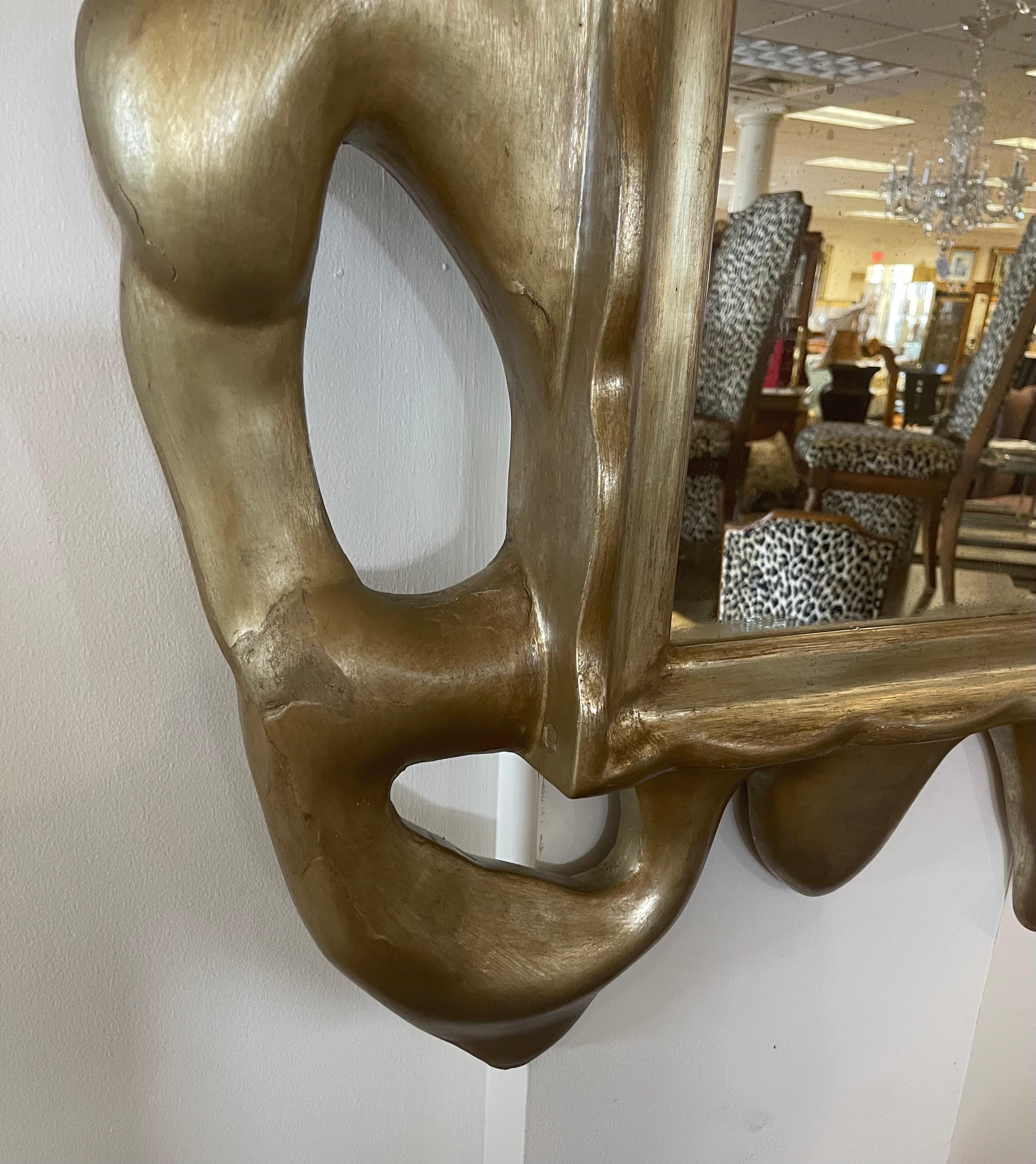 A Donghia hand carved wood frame with freeform, organic lines. With a muted lighter gold finish on the frame and antiqued patina on the mirror itself, this luxe piece hints at traditional styles but feels fresh and modern. There is some age