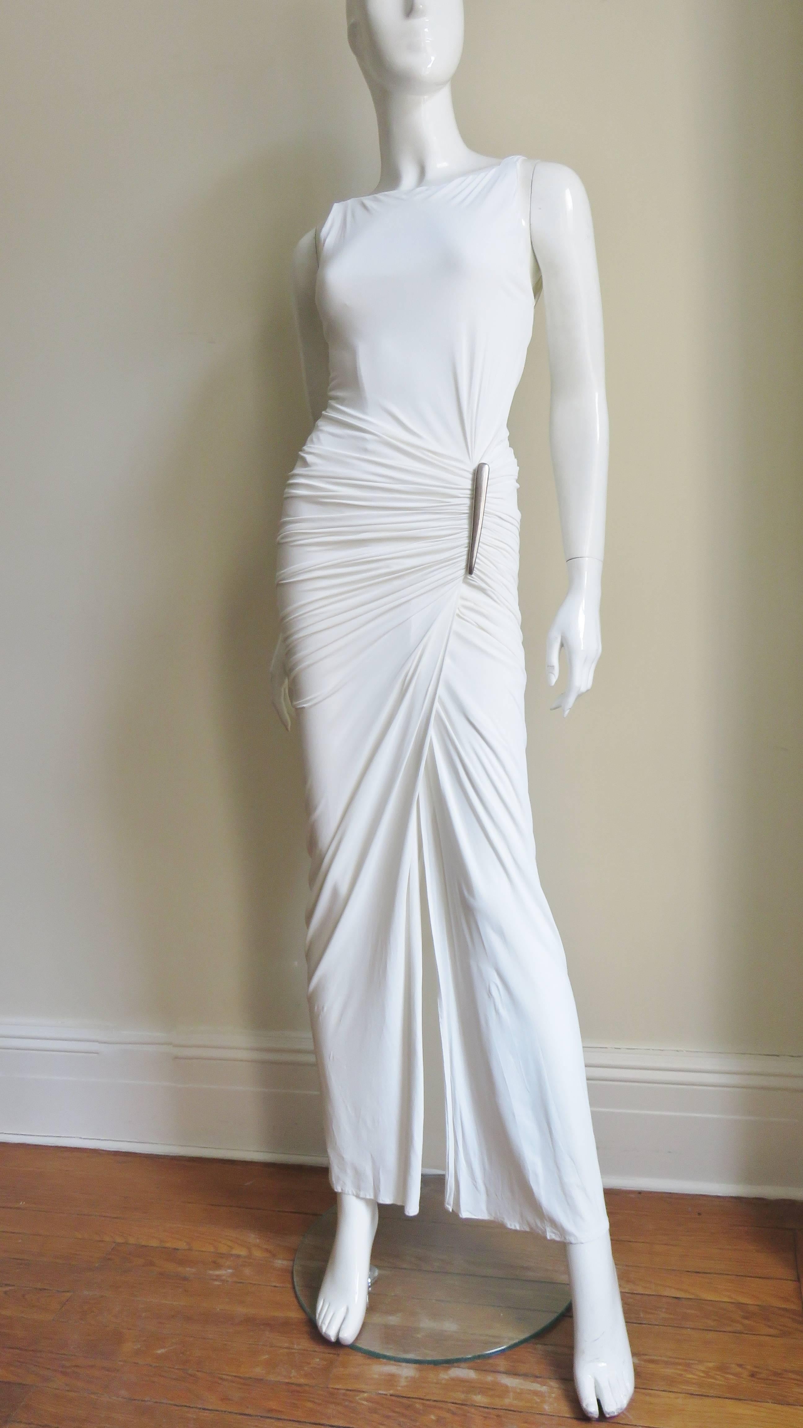 A stunning white silk jersey gown by Donna Karan.  It is sleeveless fitted with fabulous ruching across the hips accented at one side with silver metal hardware.  There is a front opening exposing a bit of leg and the upper back crosses forming a V.