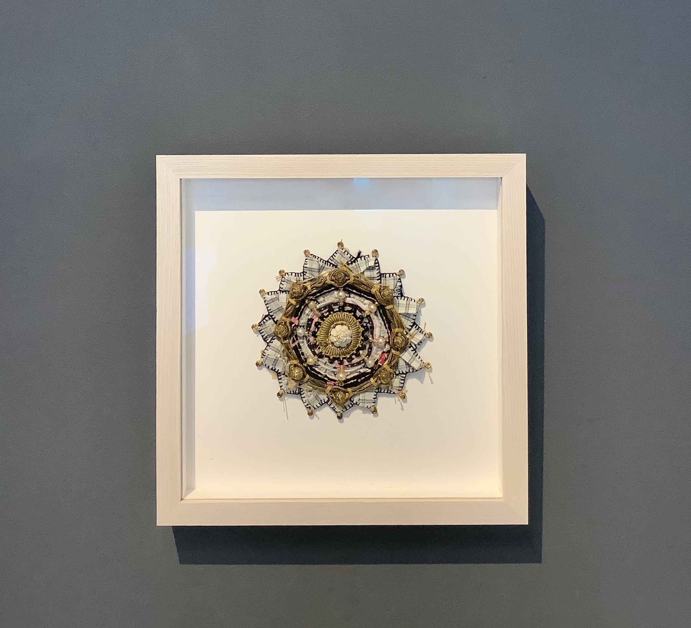 Este mandala contemporáneo de técnica mixta de Donna Sharrett se compone de puntas de cuerda de guitarra, ropa, labores de aguja, joyas e hilo. Una cuenta de rosa de marfil tallada en el centro está perfilada con hilos de oro y un círculo de perlas,