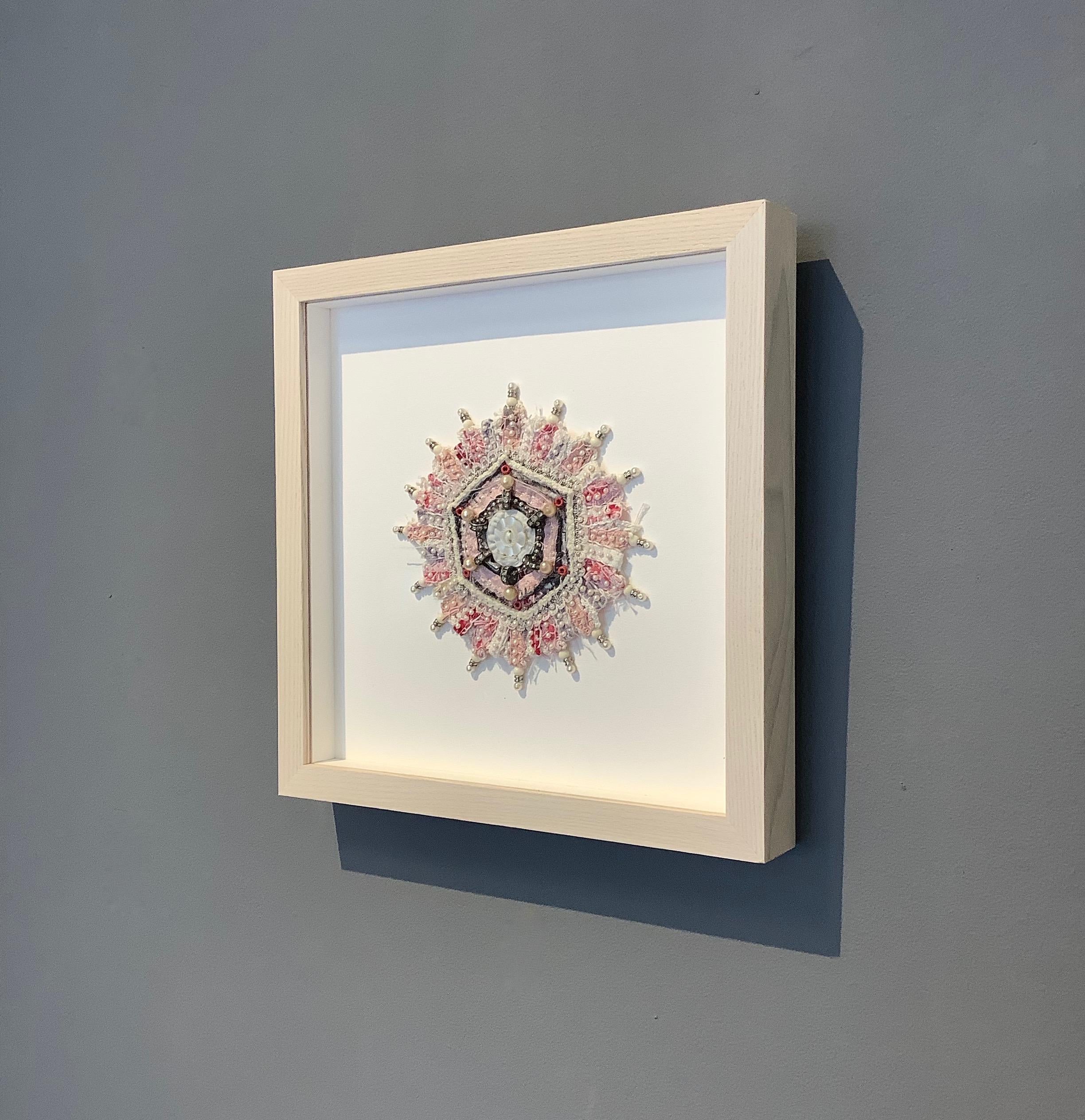 This mixed media mandala by Donna Sharrett is composed of guitar string ball-ends, clothing, needlework, jewelry, and thread, and is framed in a square 10 x 10 inch light natural wood float frame (5 x 5 inches unframed). 

Rhinestone studded beads