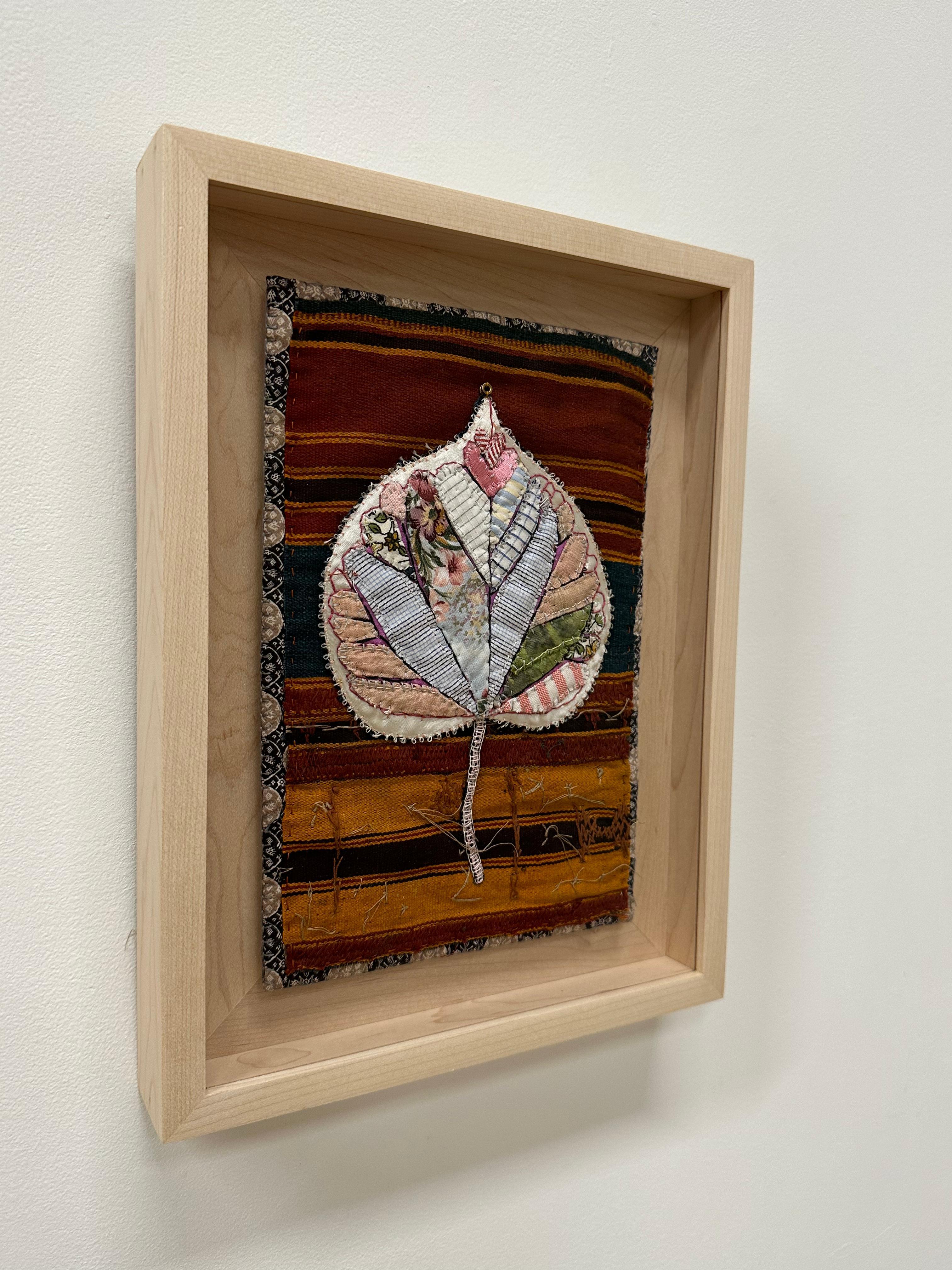 An eastern redbud leaf is composed of delicately stitched donated clothing, textiles, notions, buttons and guitar string ball-ends, in warm umber brown, saffron, light pink, sage green and a botanical pattern. 

Floated on a natural maple wood