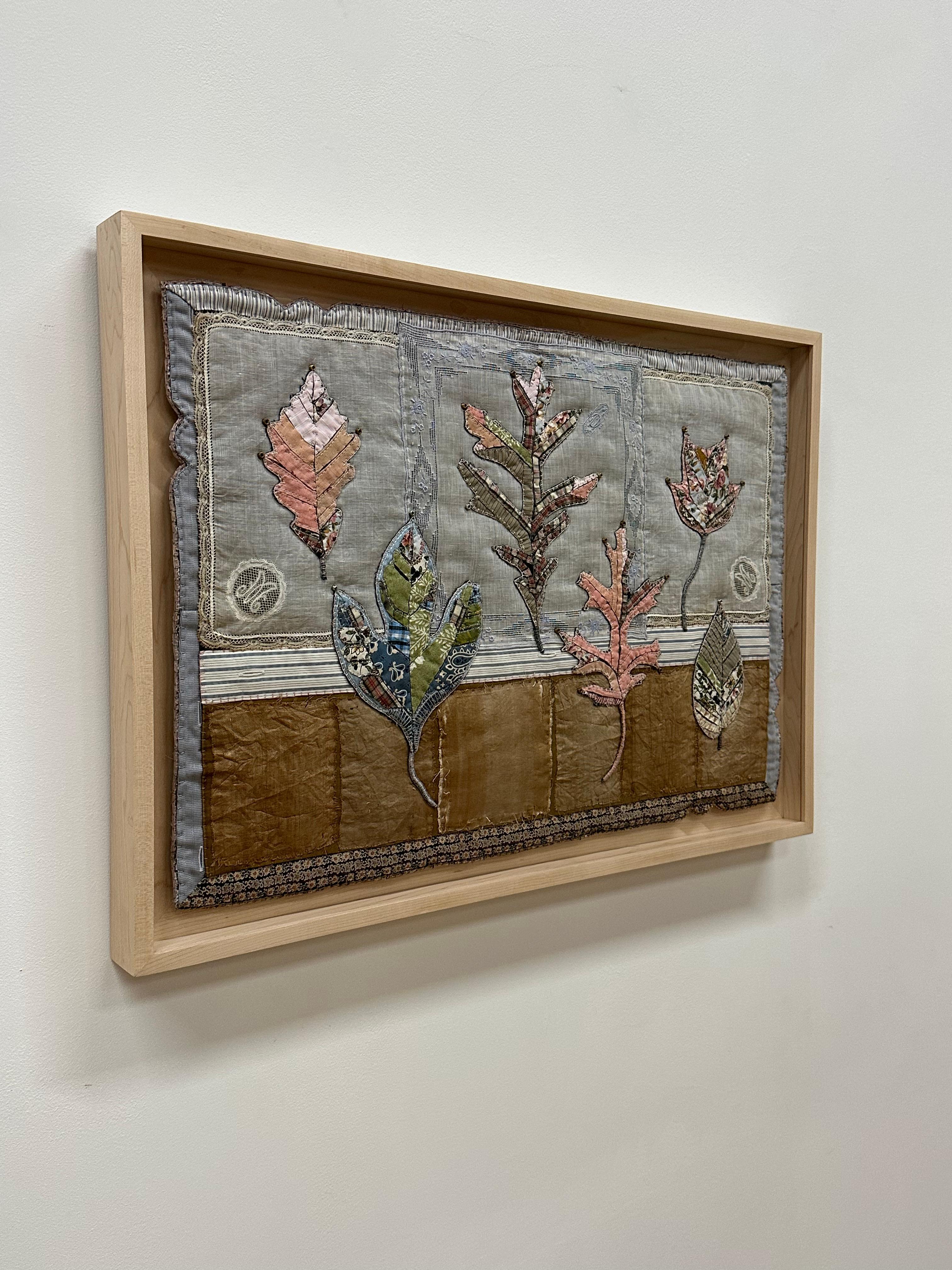 In this contemporary botanical textile art, a group of leaves in various shapes are composed of delicately stitched donated clothing, lace, notions, buttons and guitar string ball-ends, in a soft palette of pale salmon, light blue, a checkered red