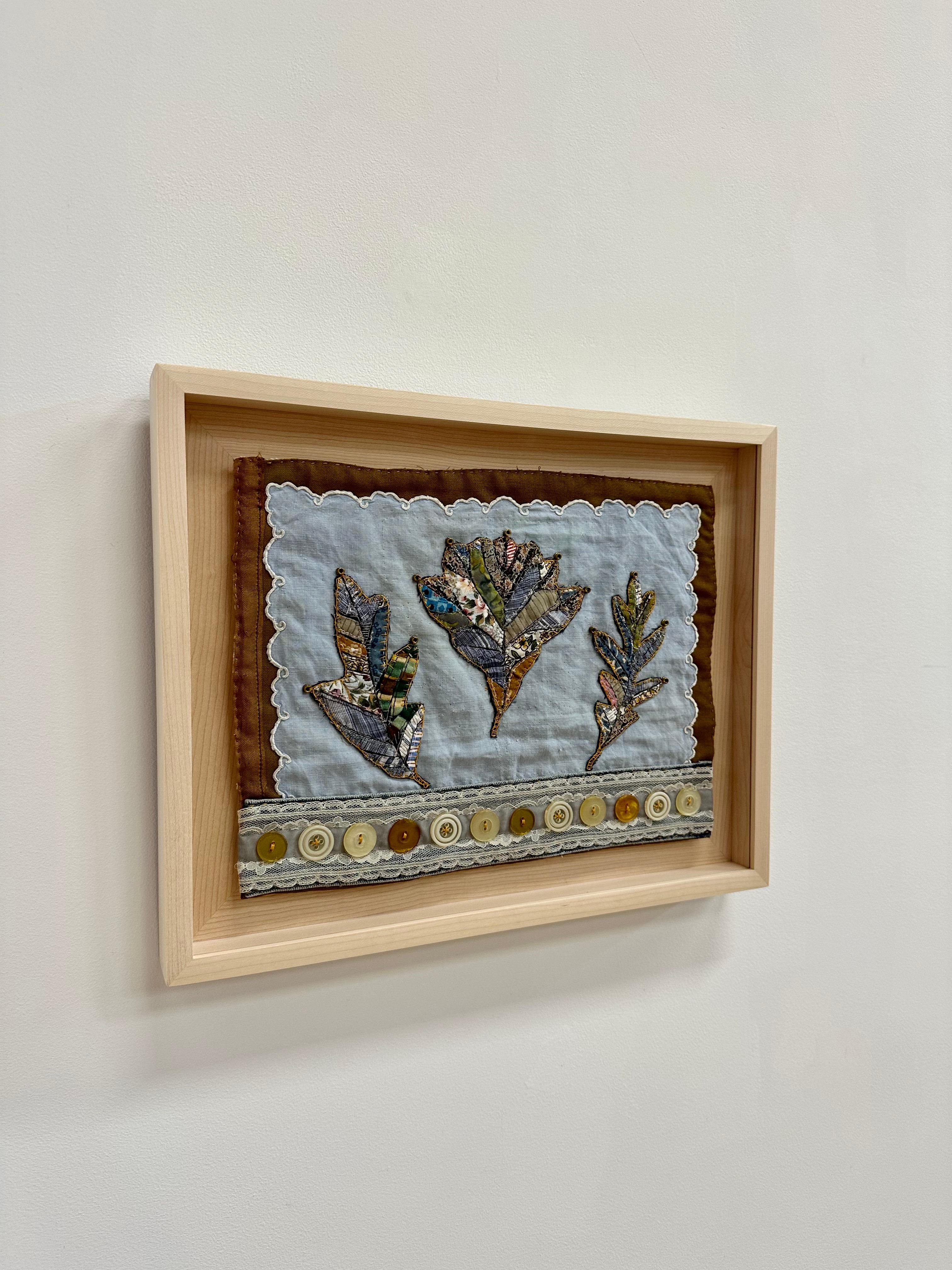 In this contemporary botanical textile, a group of leaves in various shapes are composed of delicately stitched donated clothing, textiles, notions, buttons and guitar string ball-ends, in yellow ochre, a blue checkered pattern and botanical