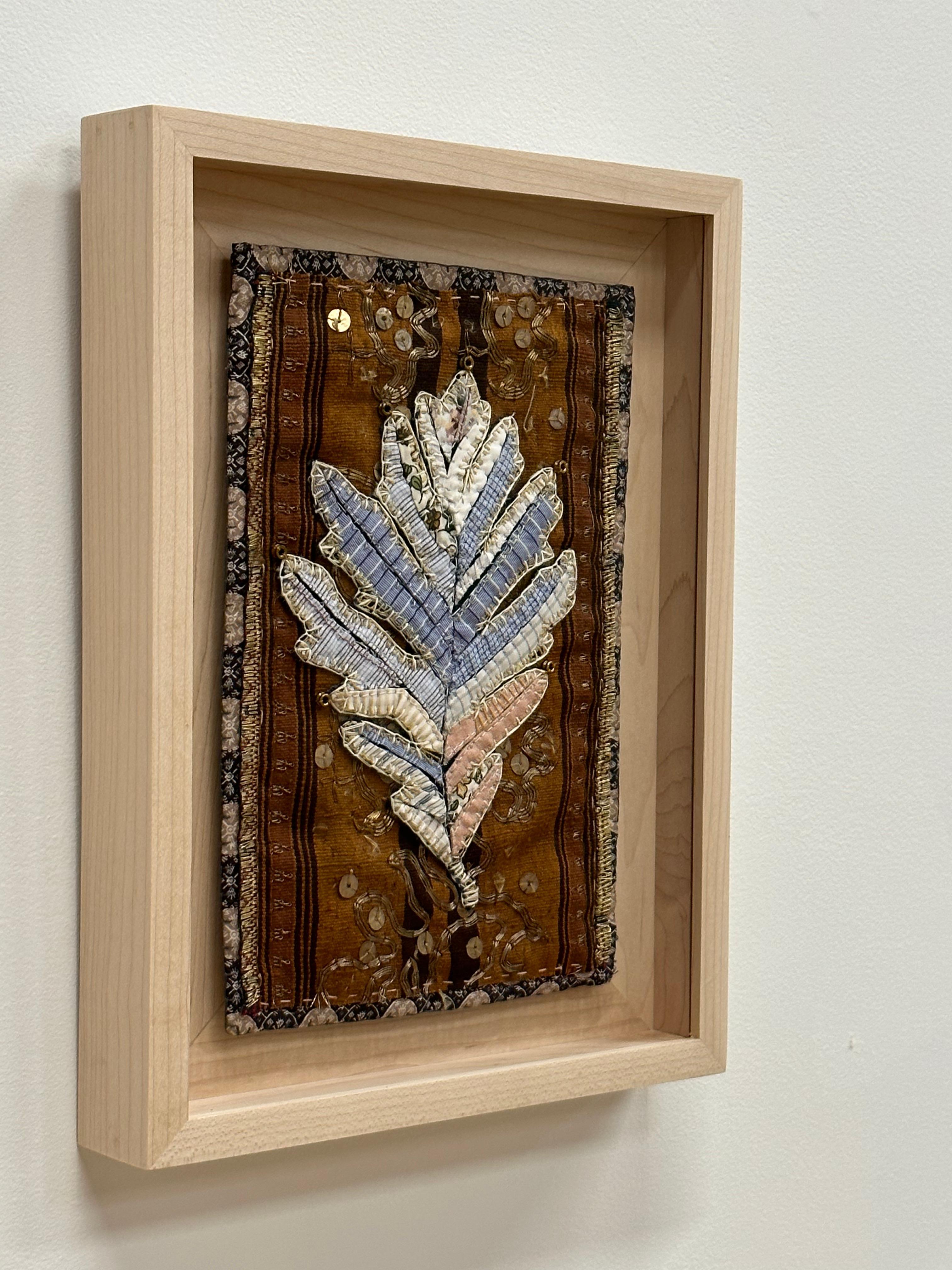 A white oak leaf is composed of delicately stitched donated clothing, textiles, notions, buttons and guitar string ball-ends and embellished with gold sequins, in warm golden brown, white, light peach, and pale blue checkered pattern and botanical