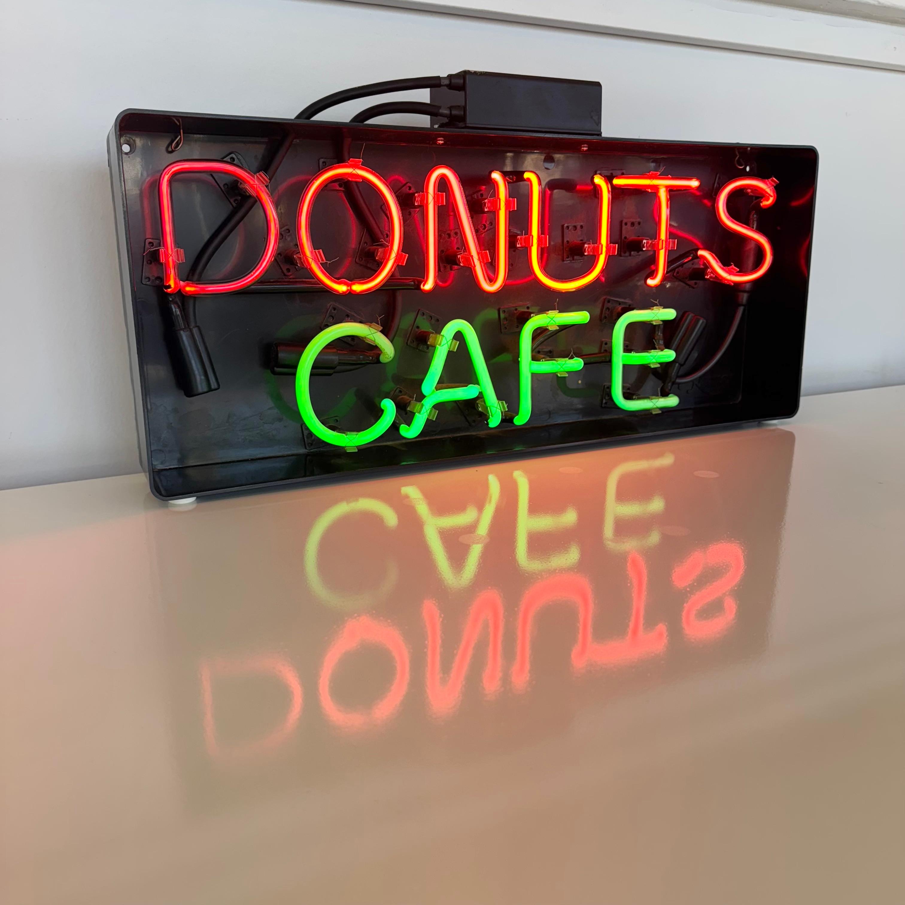 Una graziosa insegna al neon vintage con la scritta Donuts Cafe, perfetta per aggiungere un tocco nostalgico al tuo spazio. Tolta da un ristorante di Los Angeles su Hollywood Blvd. Questa piccola insegna è caratterizzata da vivaci luci al neon che