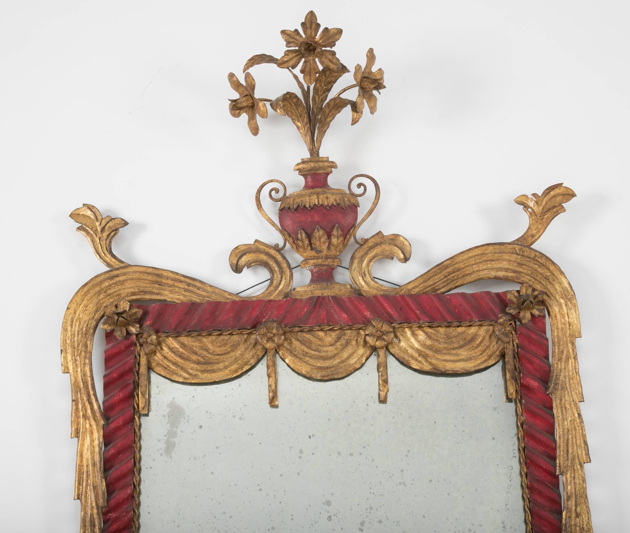 Italian Rococo style painted tole mirror, the 'Venetian Red' ribbon frame with gilt faux drapery swags topped by an urn vase of gilt metal flowers. The color and style of this mirror are very much in the Hollywood Regency style of Dorothy Draper.
