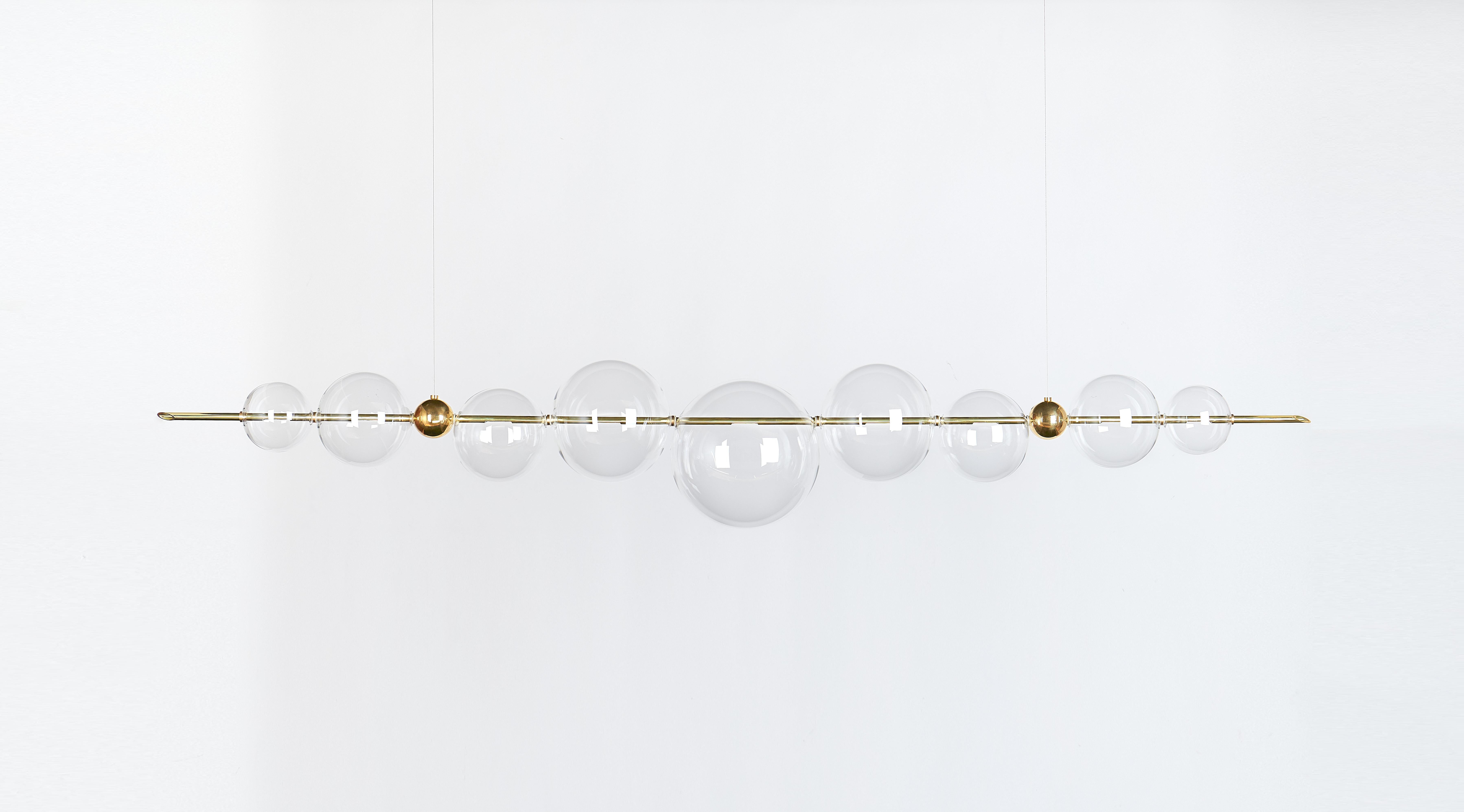 An exquisite exercise in balance, this chandelier exudes a Minimalist flair marked by clean and essential lines. Its dimmable structure features two brass bars hanging from a brass ceiling rose, and strikingly balancing a series of delicate spheres