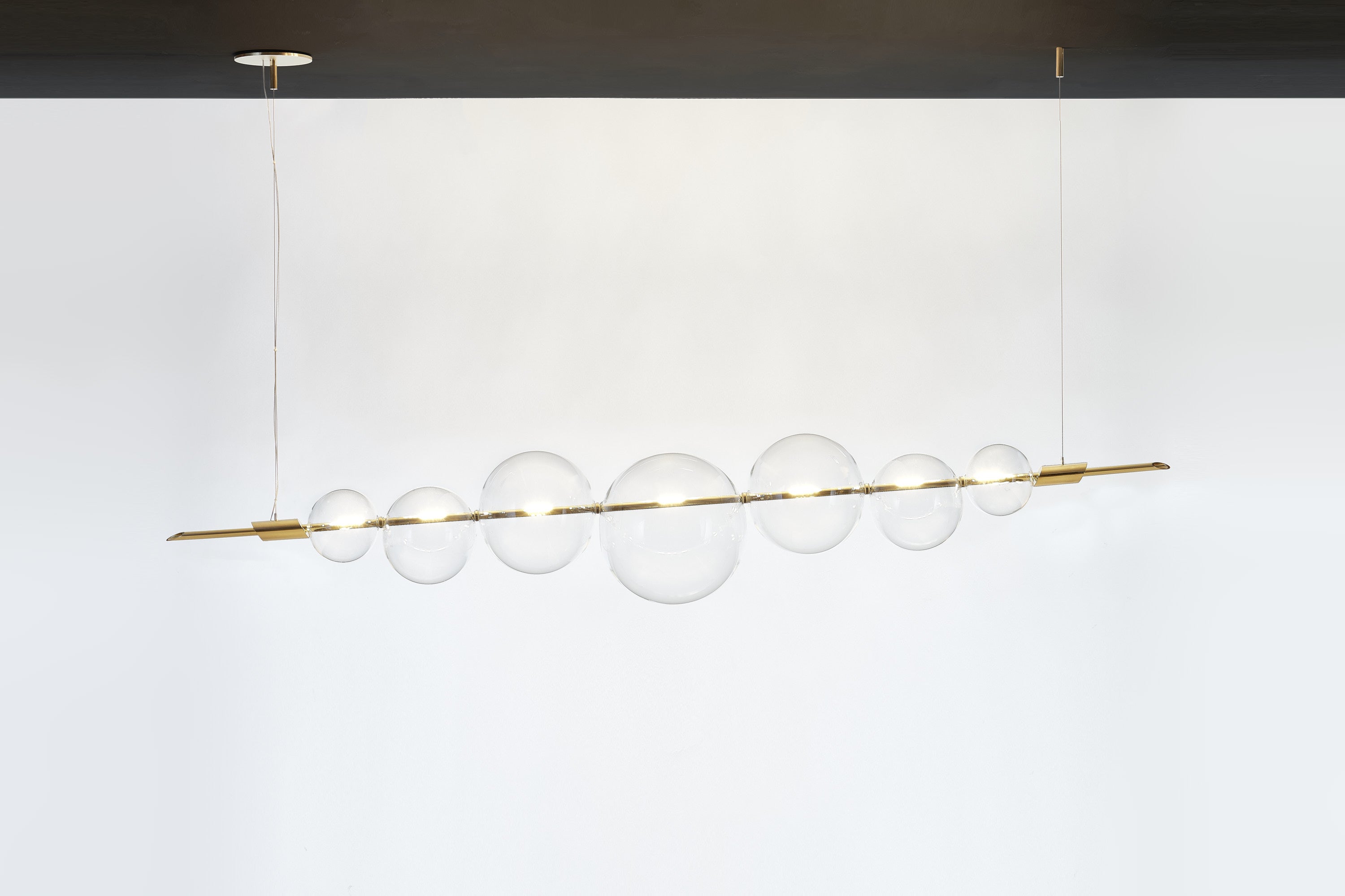 An exquisite exercise in balance, this chandelier exudes a Minimalist flair marked by clean and essential lines. Its dimmable structure features two brass bars hanging from a brass ceiling rose, and strikingly balancing a series of delicate spheres