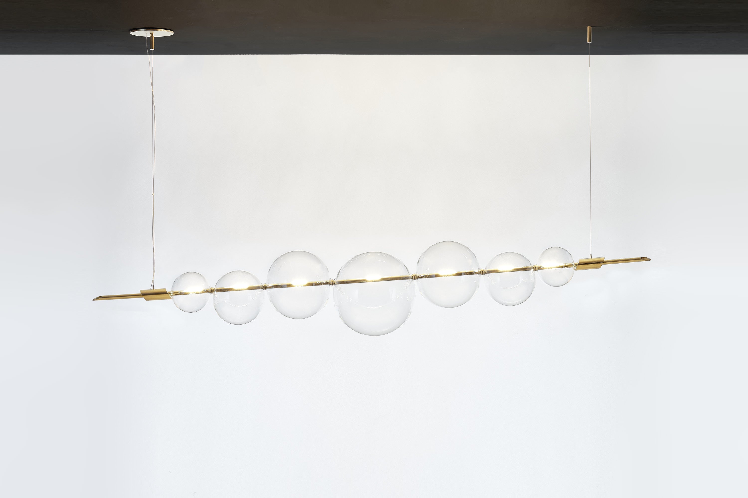 An exquisite exercise in balance, this chandelier exudes a Minimalist flair marked by clean and essential lines. Its dimmable structure features two brass bars hanging from a brass ceiling rose, and strikingly balancing a series of delicate spheres
