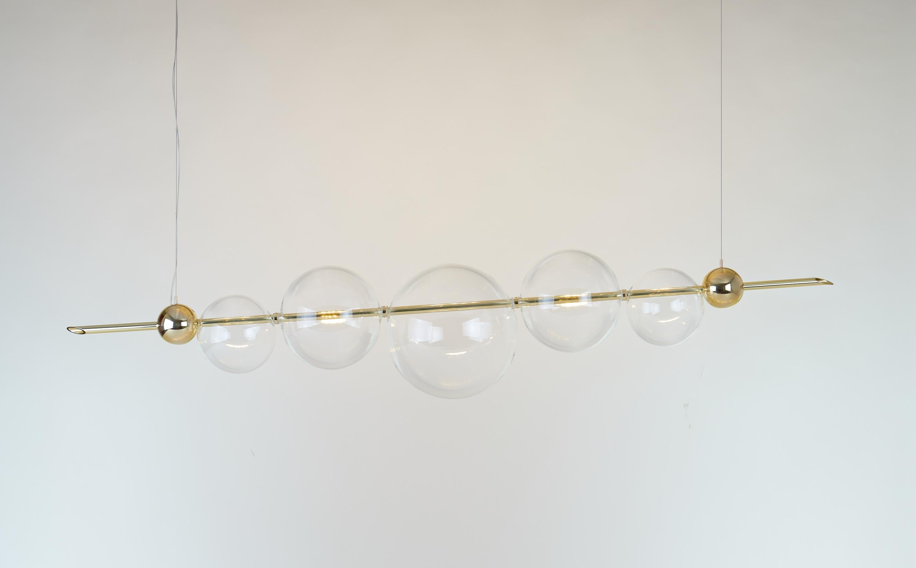 An exquisite exercise in balance, this chandelier exudes a Minimalist flair marked by clean and essential lines. Its structure features one brass bar hanging from a brass ceiling rose, and strikingly balancing a series of delicate organic globes