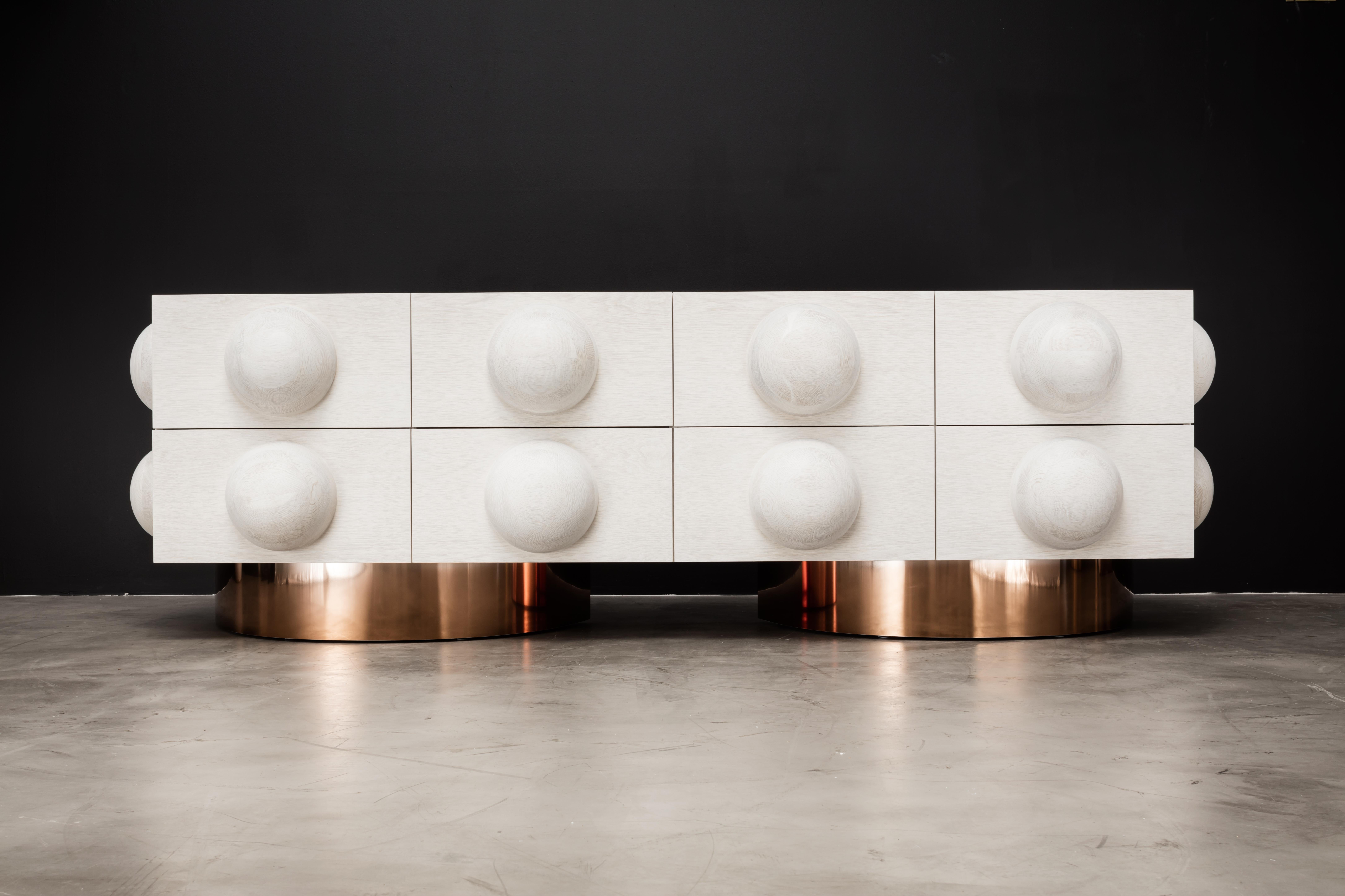 The Dot 8-drawer dresser features an architectural sculpted wood sphere detail with metal plinth bases. As shown with 6 touch latch/soft close drawers. This showroom sample is shown in bleached white oak with polished ombre bronze plinth bases. The