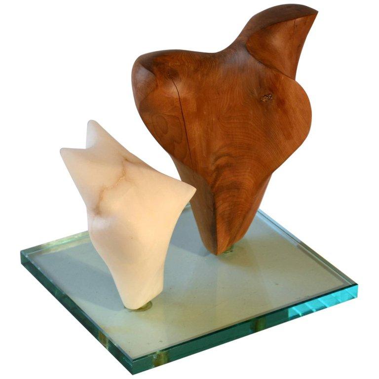 Two freeform abstract sculptures, one hand carved in wood, the other in alabaster. Together they are mounted on a square glass base.
   