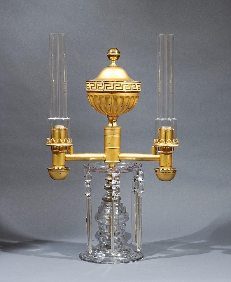 English.
Double Argand lamp, circa 1810.
Glass, blown and gilt bronze and -brass, with lamp mechanism, and with glass chimneys.
Measures: 20 1/16 in. high, 11 3/4 in. wide, 5 3/4 in. deep (overall).

The horizontally reeded blown glass base of this