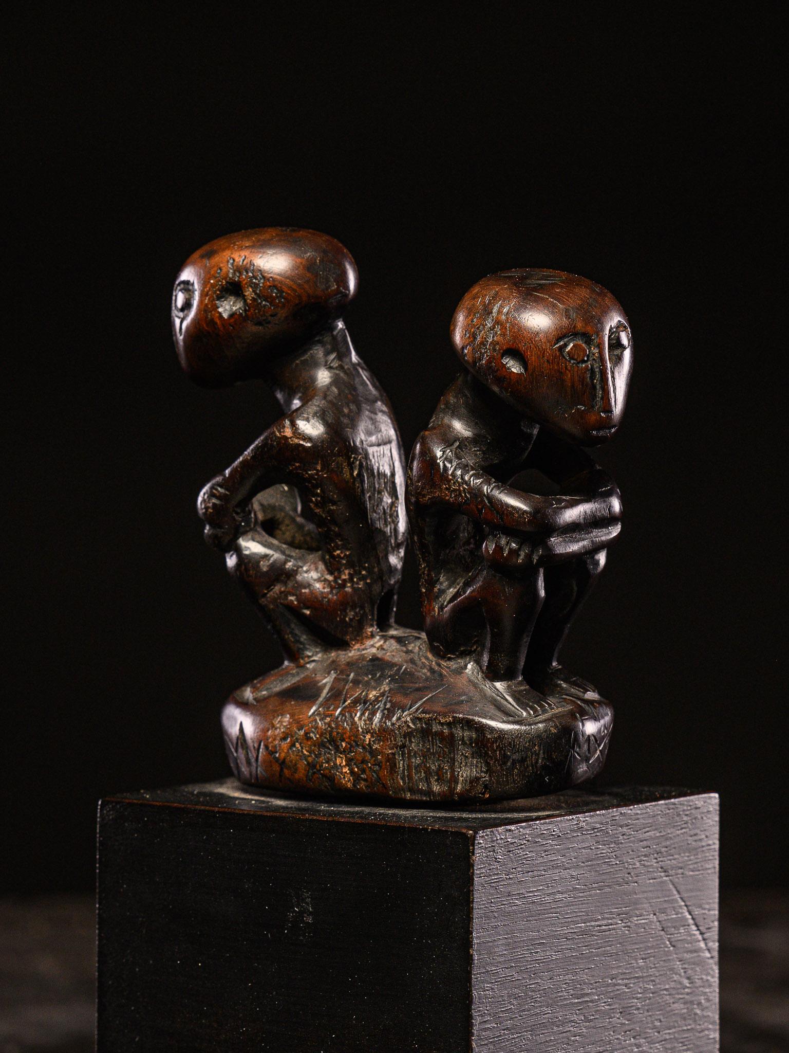 Un manico di gong in legno con due figure posizionate schiena contro schiena. Il pezzo presenta una patina liscia con una finitura scura simile al mogano. La maggior parte di queste figure sono rappresentate sedute con le ginocchia sollevate.