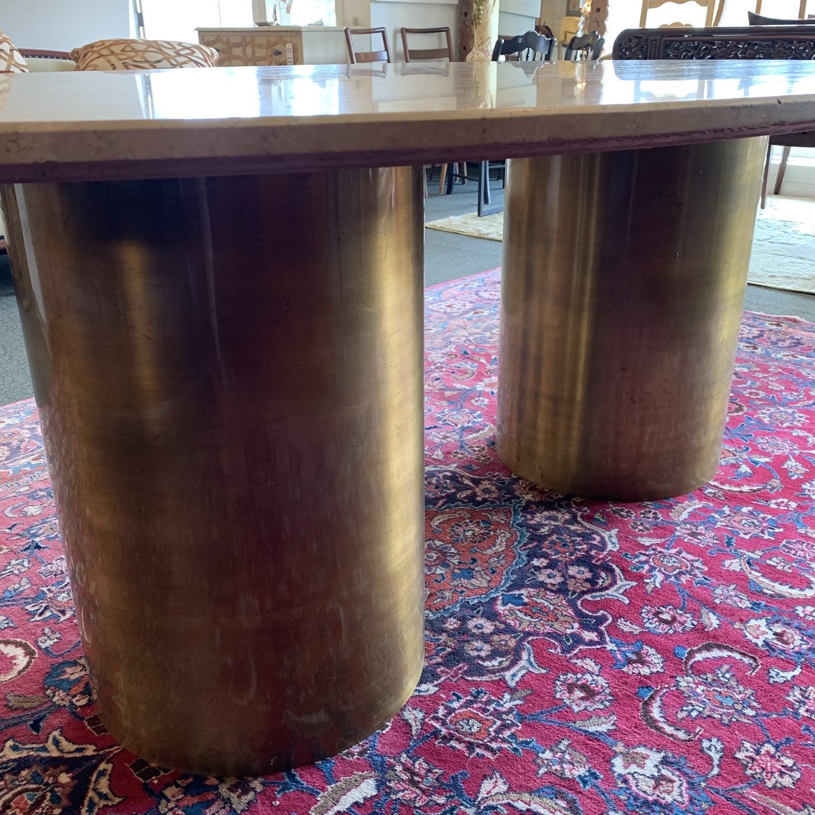 Presents a modern dining table with an 1980s vibe. A pair of spun brass columns support a lovely oval of travertine, covered in rippling waves of tan and white. The beveled edge slab is large enough to seat eight comfortably. Evidence of wear on