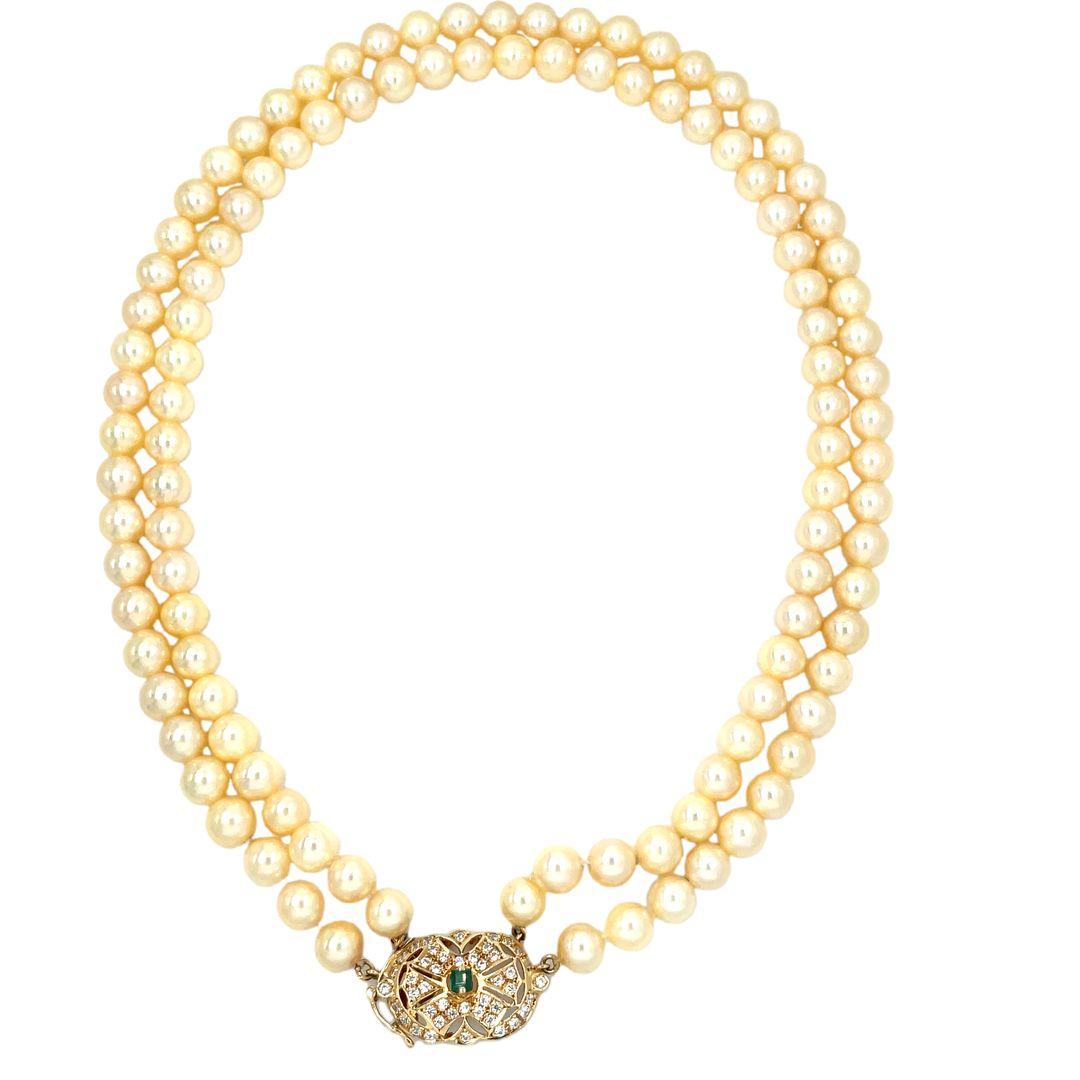 A classic double strand pearl necklace. Composed of two strands of well matched cream colored cultured pearls and completed by a 14 karat yellow gold and diamond clasp highlighting a square shaped emerald of approximately 0.30 carat. Total diamond
