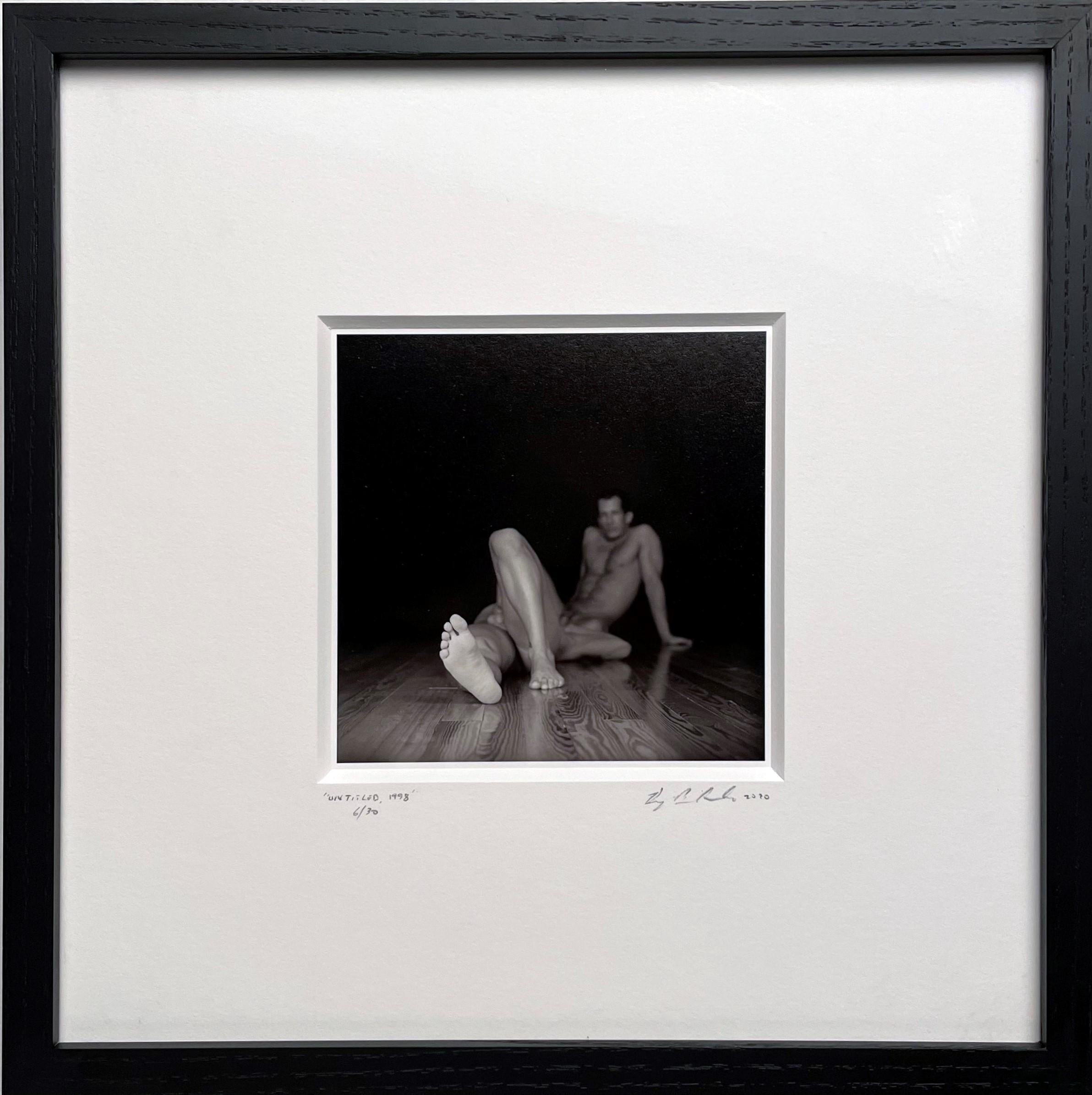Two nude males lie on a bare wood floor.  The angle of the photograph suggests that their bodies have melded into one.  The artist uses traditional photography methods shooting the image on film.  The image is printed using traditional darkroom