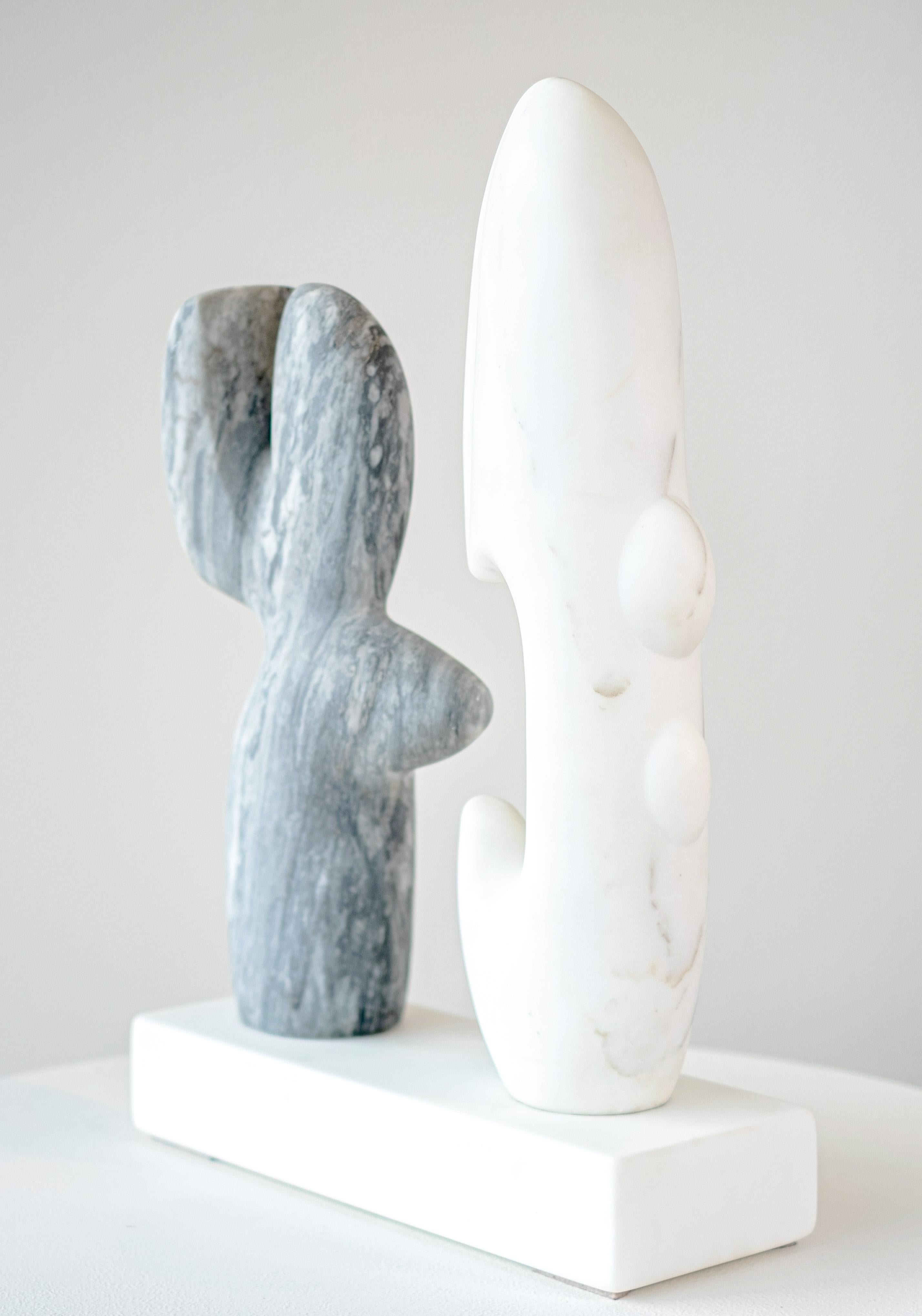 This contemporary, tabletop, white, grey, abstract, marble sculpture is by Canadian artist Doug Robinson. Two figurative shapes—one hand sculpted from creamy white Carrera marble; the other from gray Bardiglio marble appear to be having ‘a