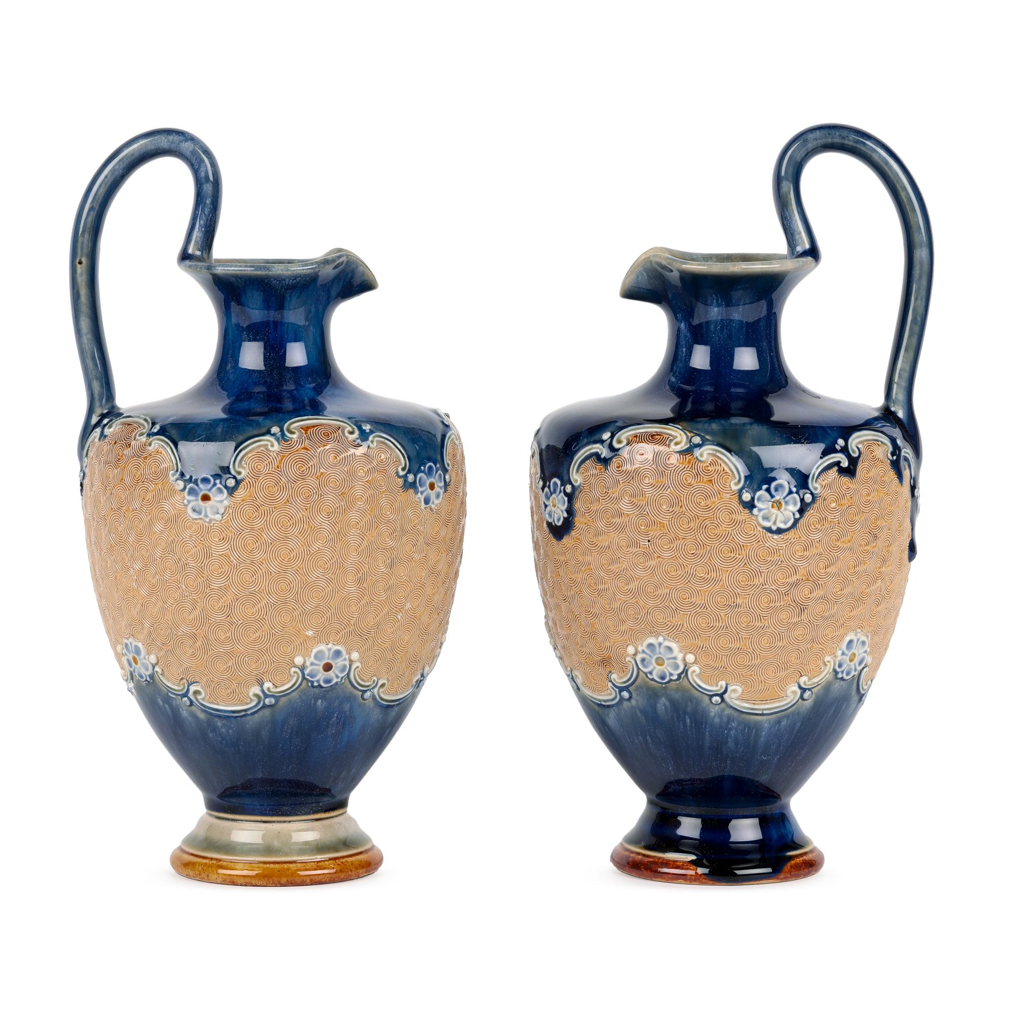 Fino par de aguamaniles de cerámica art nouveau Doulton Lambeth decorados con motivos florales y volutas, posiblemente del célebre artista John Broad, de principios del siglo XX. Los aguamaniles de gres, de formas elegantes, se elevan sobre un