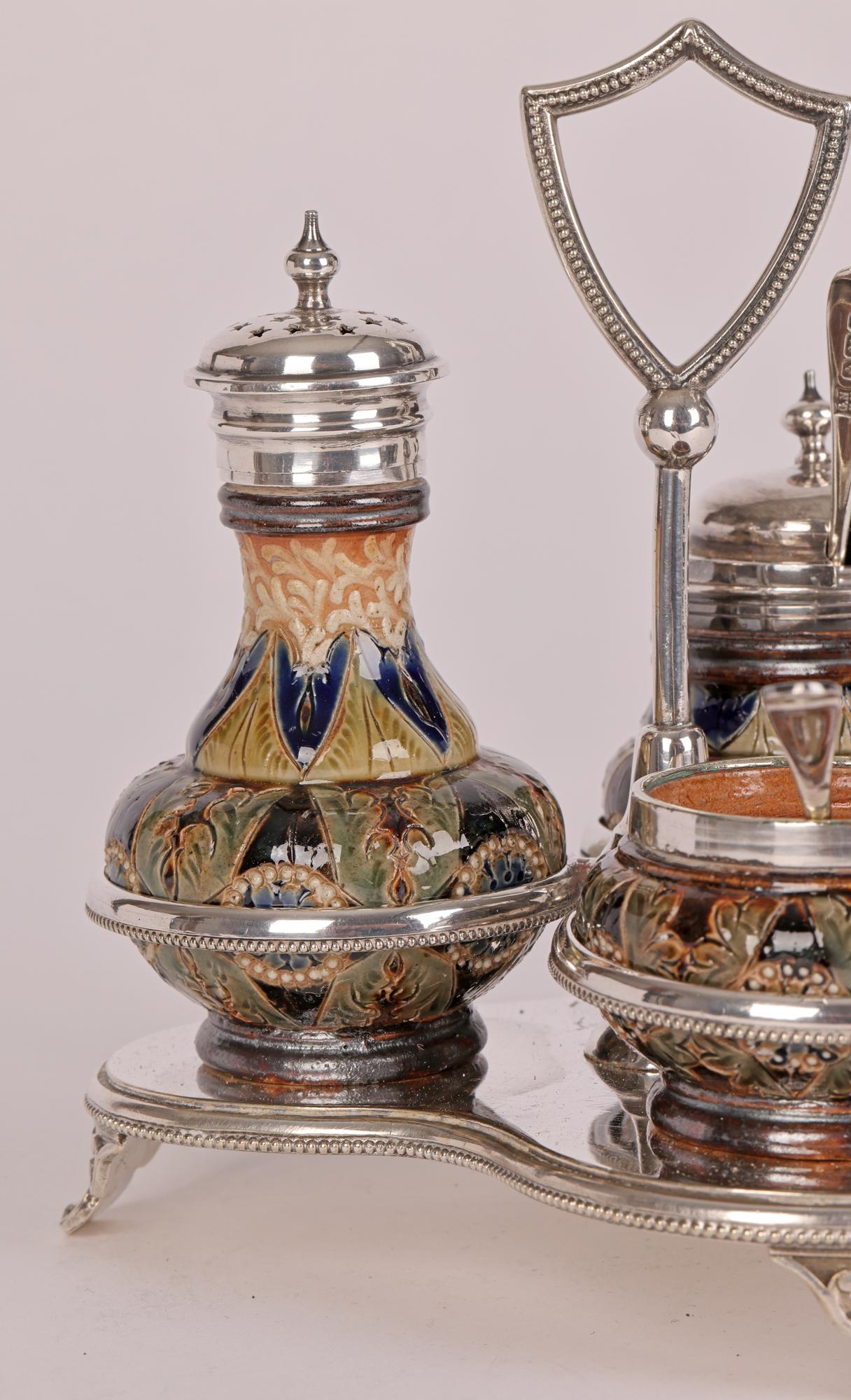 A very fine and rare Doulton Lambeth silver-plate mounted three piece stoneware cruet set contained in a decorative silver plated stand with spoons and dating from 1881. The cruet set comprises of an open salt with salt spoon, a pepper pot and a