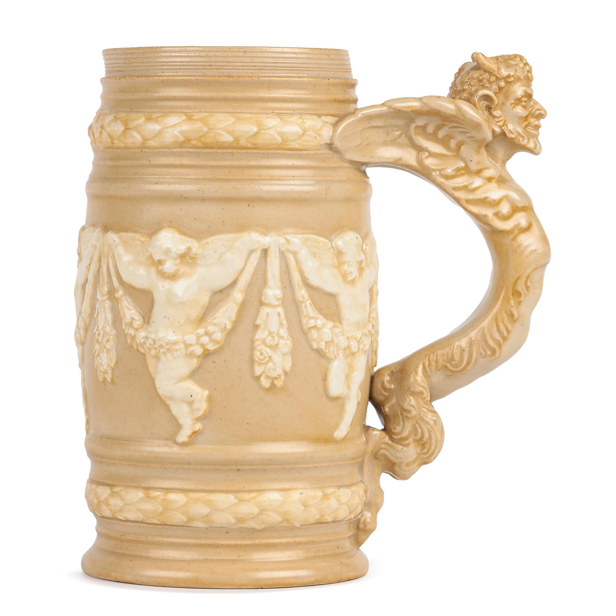 Rara e inusual jarra de cerámica artística Doulton Lambeth con asa de sátiro alado que data de alrededor de 1895. La jarra de gres es de forma cilíndrica alta y ahusada, y se alza sobre un borde de pie ligeramente más ancho con la base rebajada. El