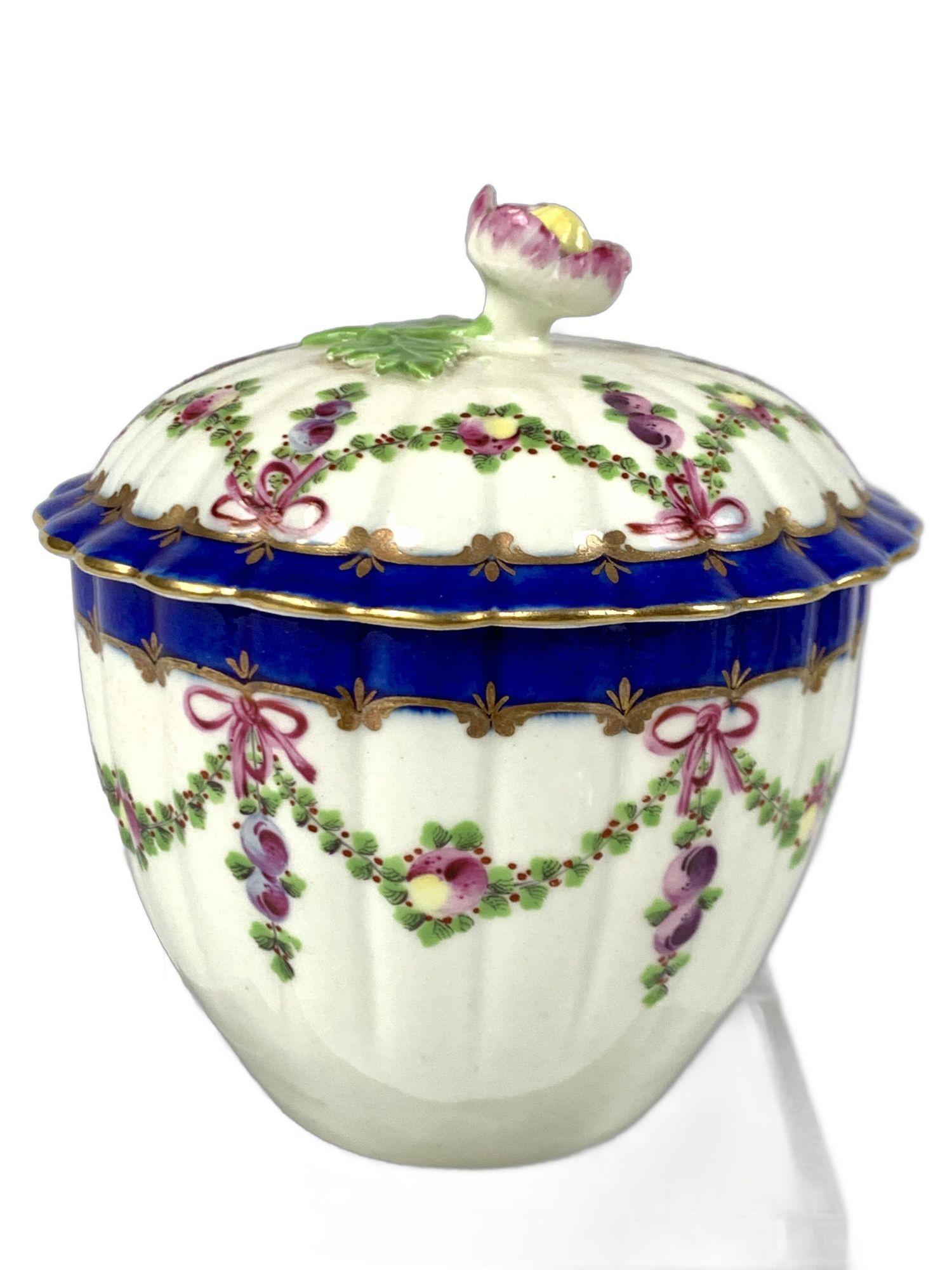 This 18th-century First Period Worcester Porcelain sugar box was hand painted featuring beautiful swags painted with green leaves, purple berries, and two-tone blue and yellow apples.
Each swag is tied with a purple ribbon in an elegant bow. Green