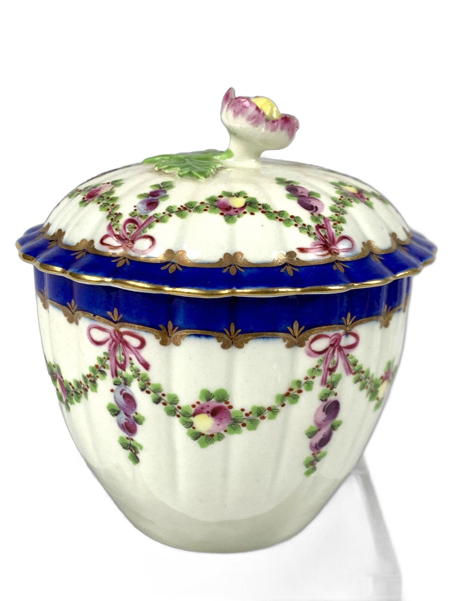 Este azucarero de porcelana Worcester de la Primera Época del siglo XVIII fue pintado a mano con hermosas guirnaldas pintadas con hojas verdes, bayas moradas y manzanas bicolores en azul y amarillo.
Cada swag se ata con una cinta morada en un
