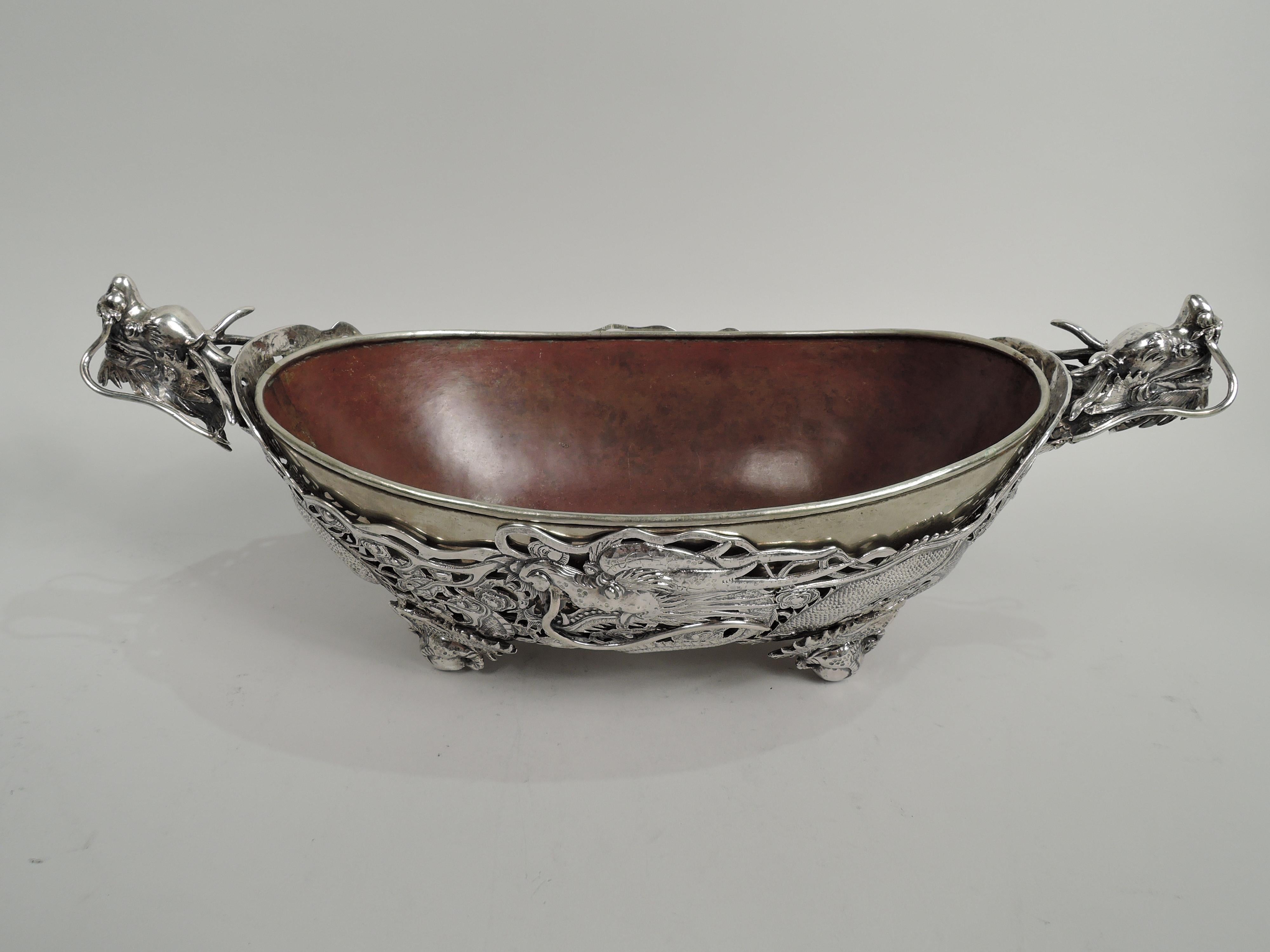 Dramatic Chinese silver centerpiece bowl, ca 1890. Oval with solid well. Sides open with scaly, slithering wraparound dragons; haunches and legs terminating in talon-gripped balls form supports. Cast heads with gaping jaws and lolling tongue.