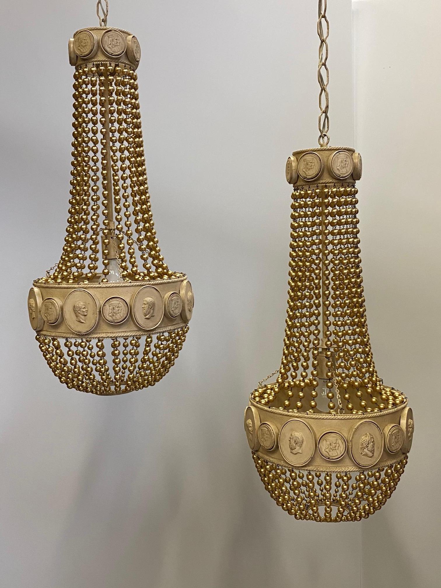 Dramatic pair of Hollywood Regency pendant chandeliers having taupe colored cast metal construction with cascading necklaces of pearl finished beads and neoclassical style medallions around the top and lower band.