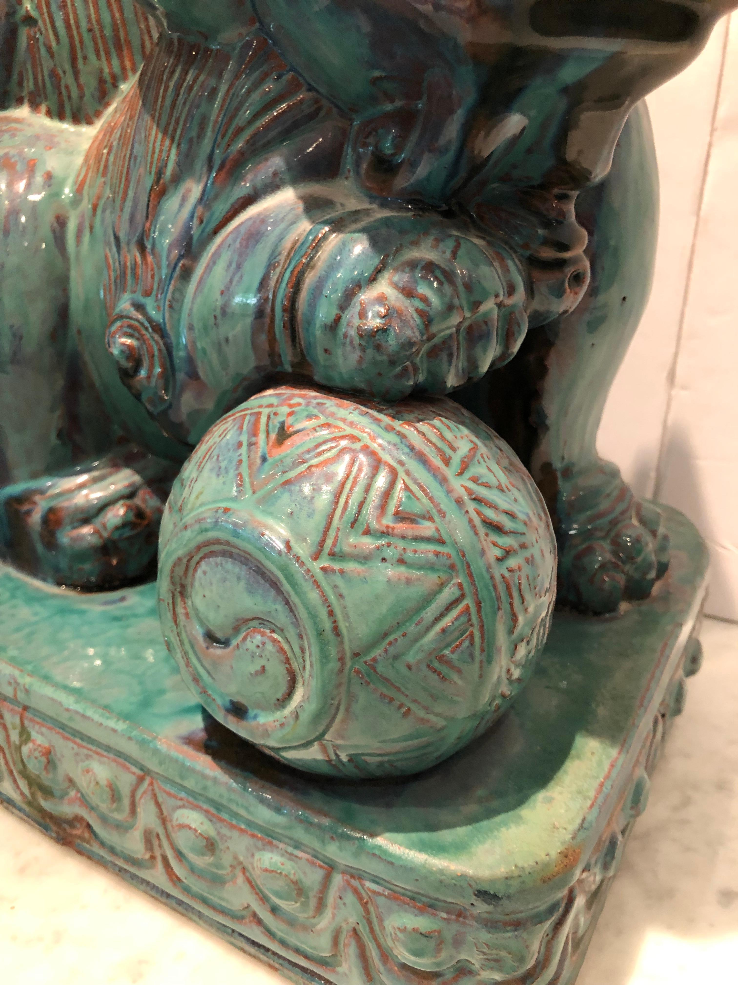 Large pair of Chinese foo dogs in a turquoise glaze. One dog has a ball under its paw, the other a smaller dog. Made of terra cotta, the chunky size of this pair make them quite dramatic. The glaze on the front of the plinth of one dog reads green