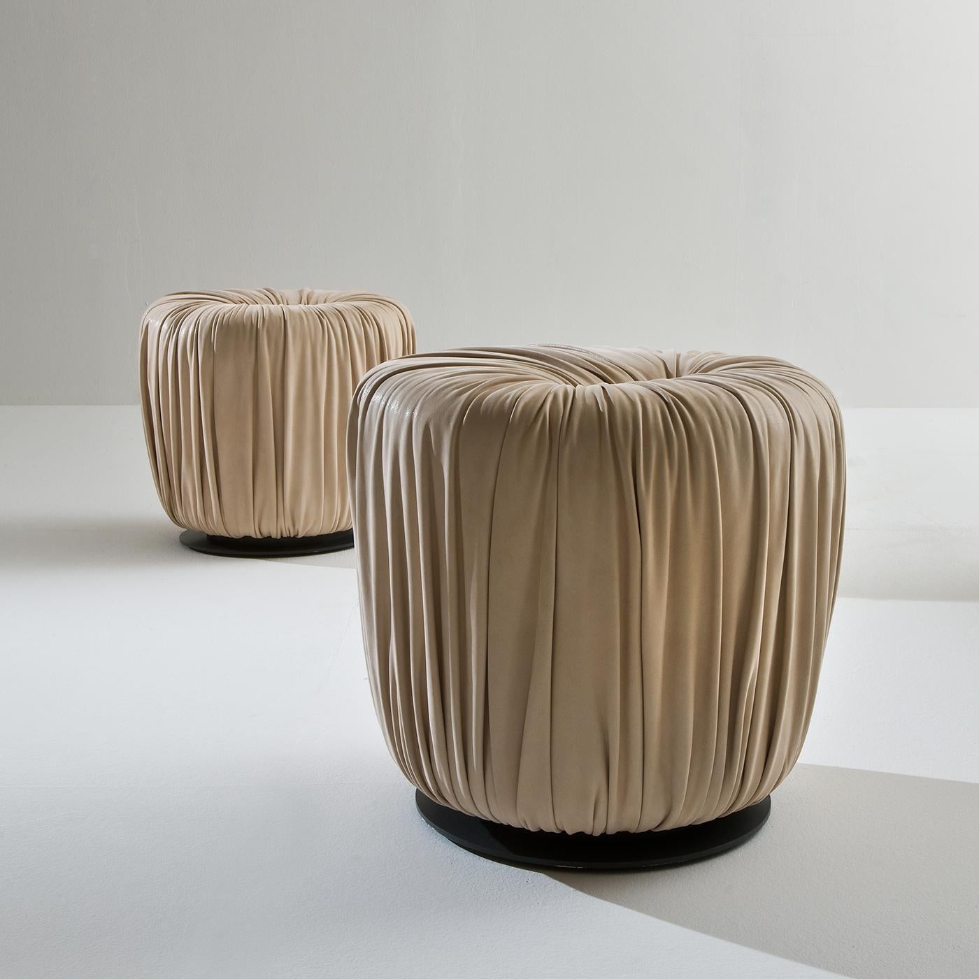 The unique sartorial work of the this pouf series is immediately recognizable for creating soft shapes. The Drapé Collection is a set of upholstered poufs covered in leather or velvet, with a base in metal. This luxurious round pouf is upholstered