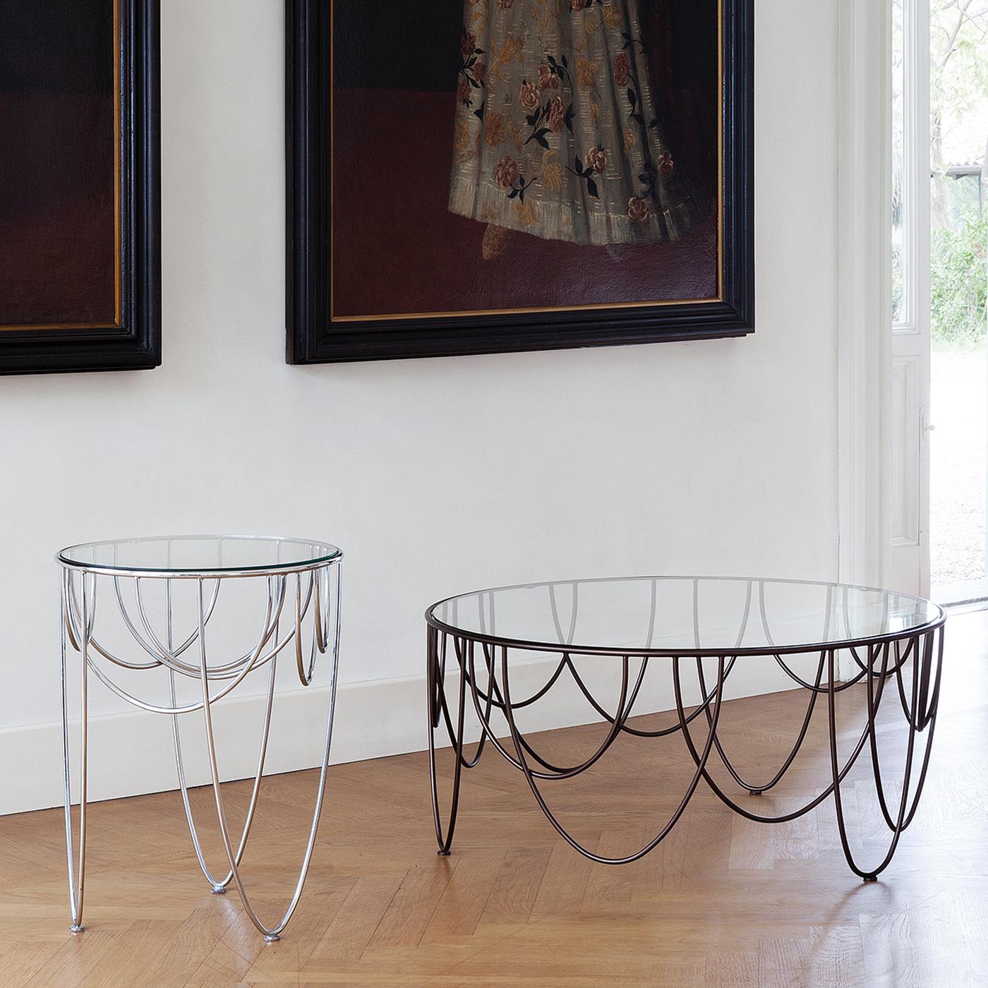 This spectacular, sculptural side table is distinguished by a captivating structure that draws inspiration from the drapery of stage curtains. Made of powder-coated steel rods, it supports a large round top in tempered crystal. This piece will