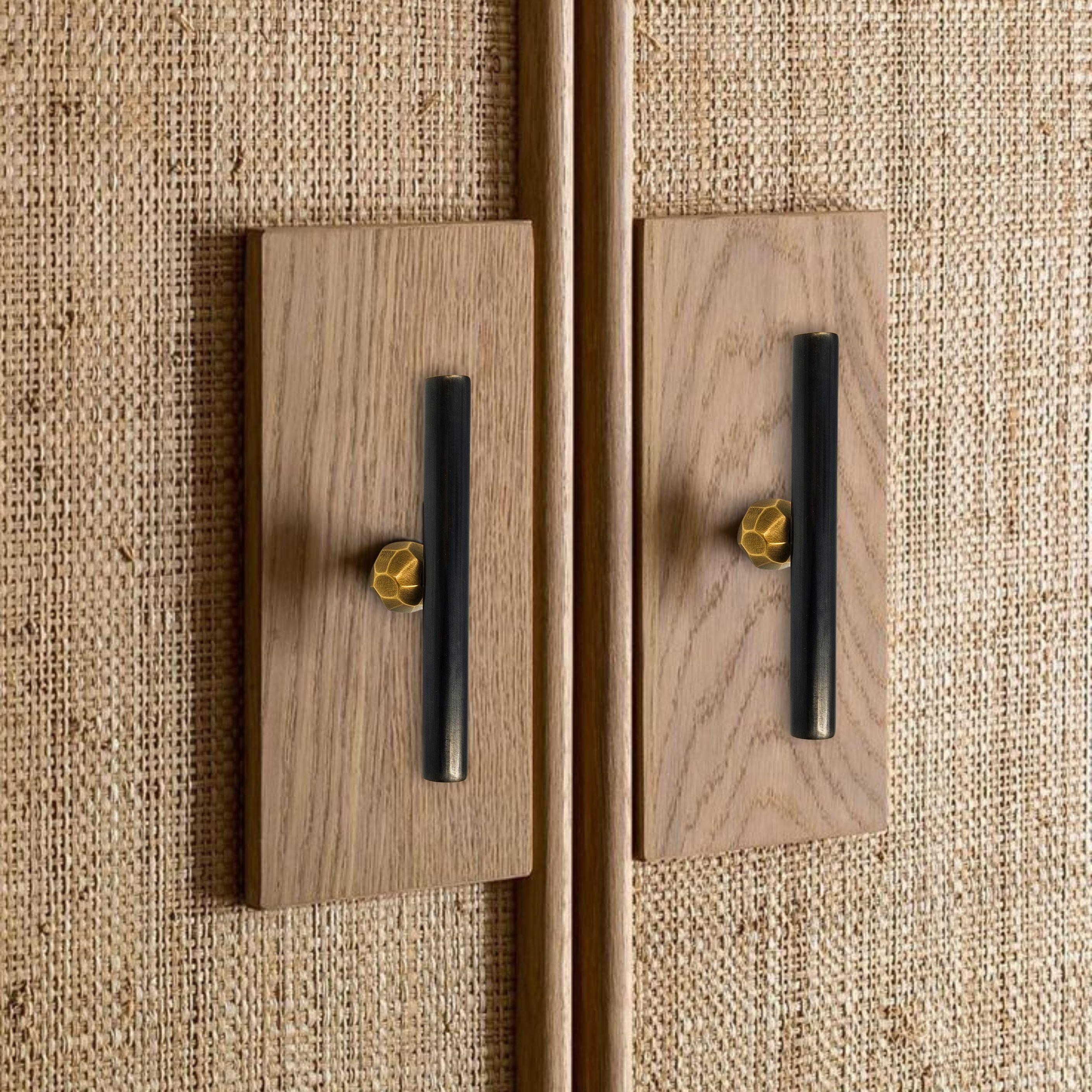 This drawer pull features a linear structure composed of a brass tube in an aged gold finish, accented at the center by a hammered button in gunmetal-patinated brass. Measuring 10 cm in length, its clean, horizontal form is enhanced by the contrast