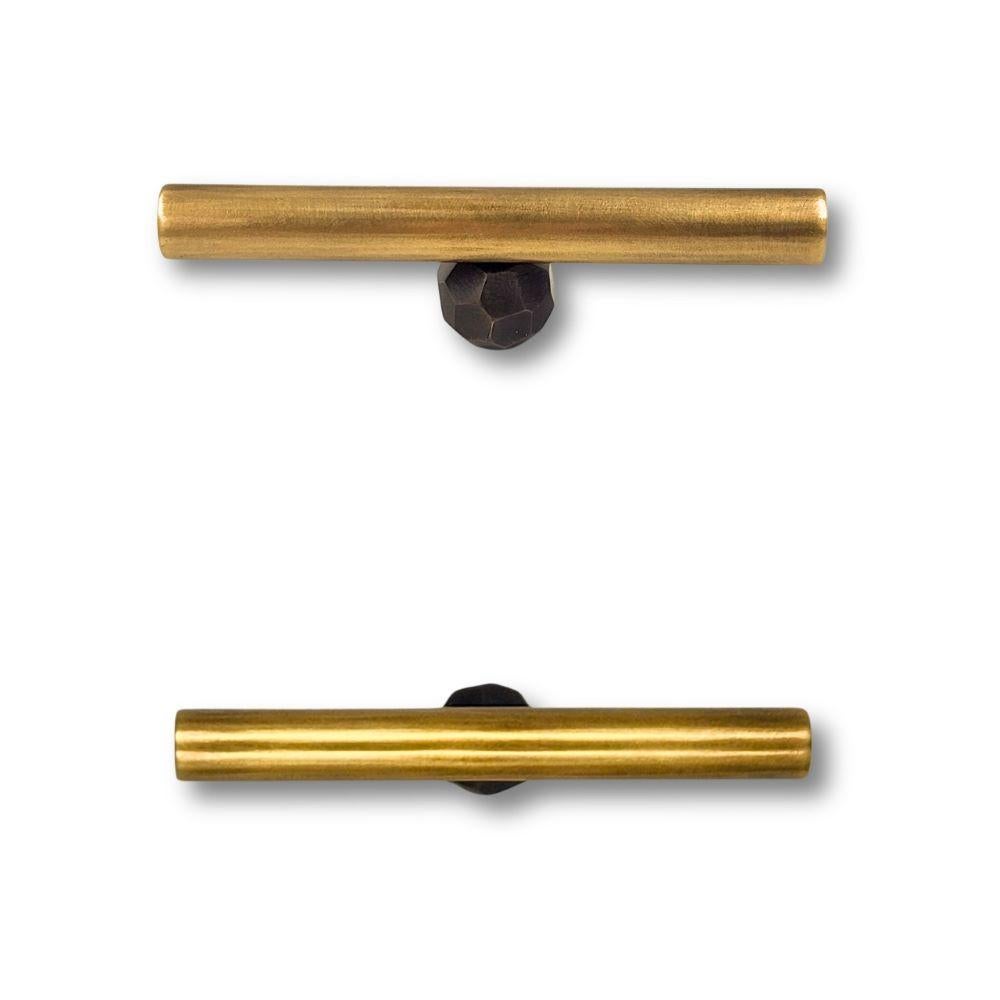 This drawer pull combines a gunmetal-patinated brass tube with a central hammered button in an aged gold finish. Measuring 10 cm in length, it features a clean, horizontal silhouette. The contrast between the dark, smooth tube and the irregular