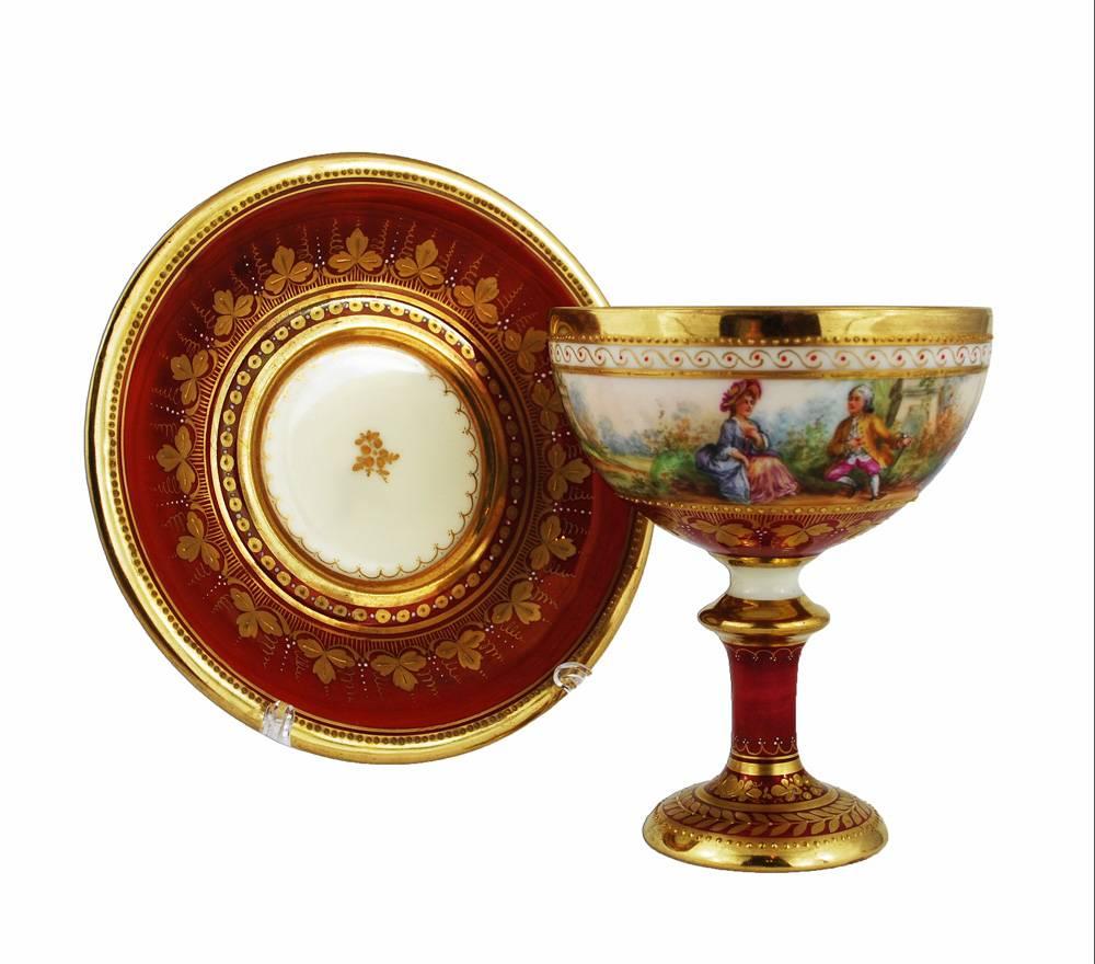 Cup has a hairline.

A hand-painted vintage footed cup and saucer from Ambrosius Lamm Studio in Dresden, circa 1930. Expertly painted scenes of courtship decorate the outside of the cup, red glaze graces the inside and stem. Heavily gilt