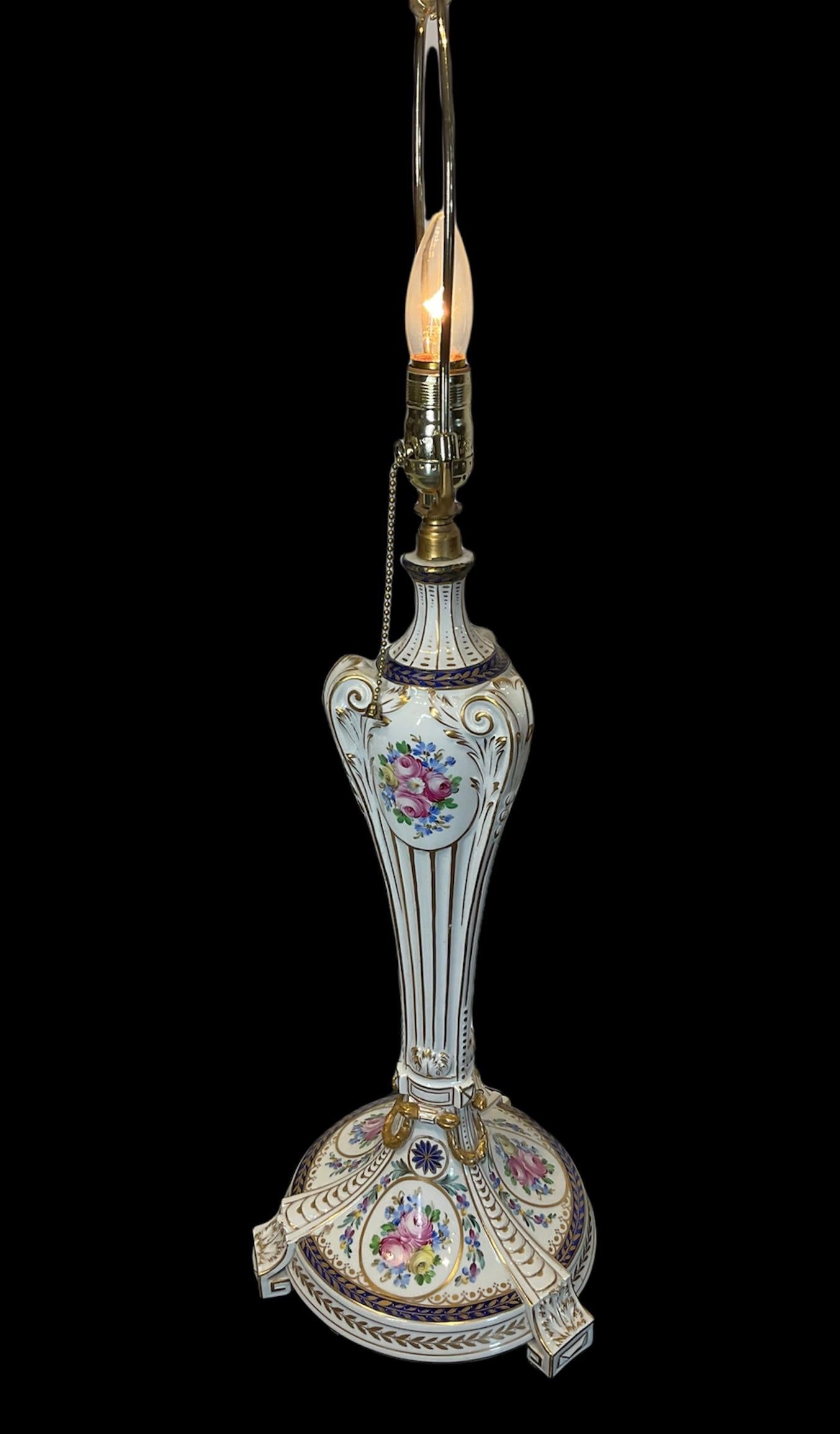 This is a Dresden hand painted porcelain table lamp. It is long and urn shaped. It has a white background with gilt fluted details that form scrolls at the top. The lamp is embellished by oval frames made of acanthus leaves with bouquets of roses in