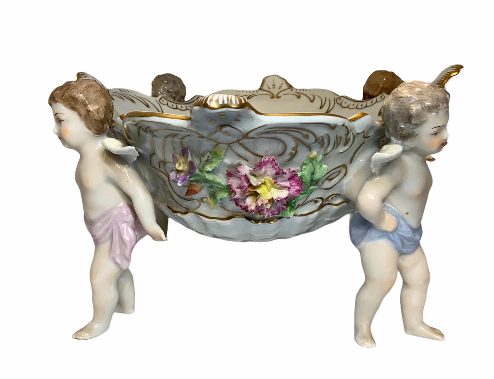 This is a Dresden porcelain depicting four winged cherubs holding a quatrefoil shaped compote. Repousse colorful flowers and gilt scrolls adorned the outside of the white compote. Inside of the compote there is hand painted bouquet of flowers and