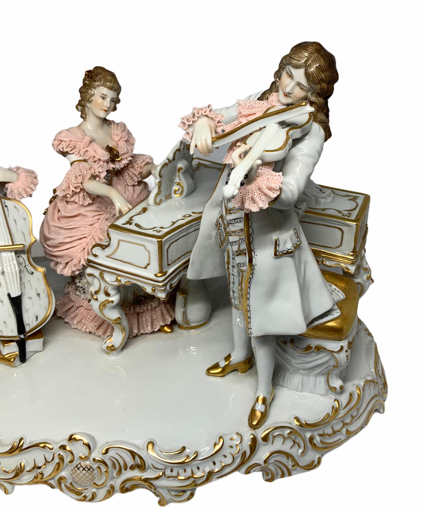 This a Dresden Porcelain group figurines depicting a trio of musicians. The group is made of a lady & two gentlemen. The lady who play the piano is wearing a delicate peach color Lace and white dress. There is an evening white lady cape over the