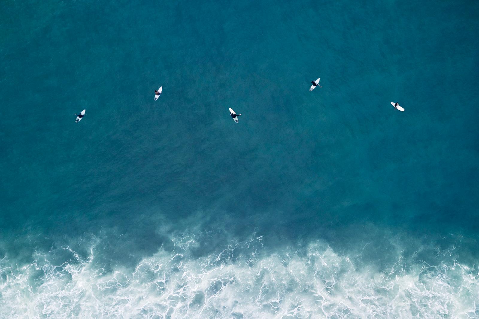 Cinco surfistas esperan la próxima serie de olas en medio del mar cerúleo - Photograph de Drew Doggett